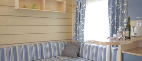 A striped sofa with a cushion, a small table with a bottle and glasses, and a window with curtains.
