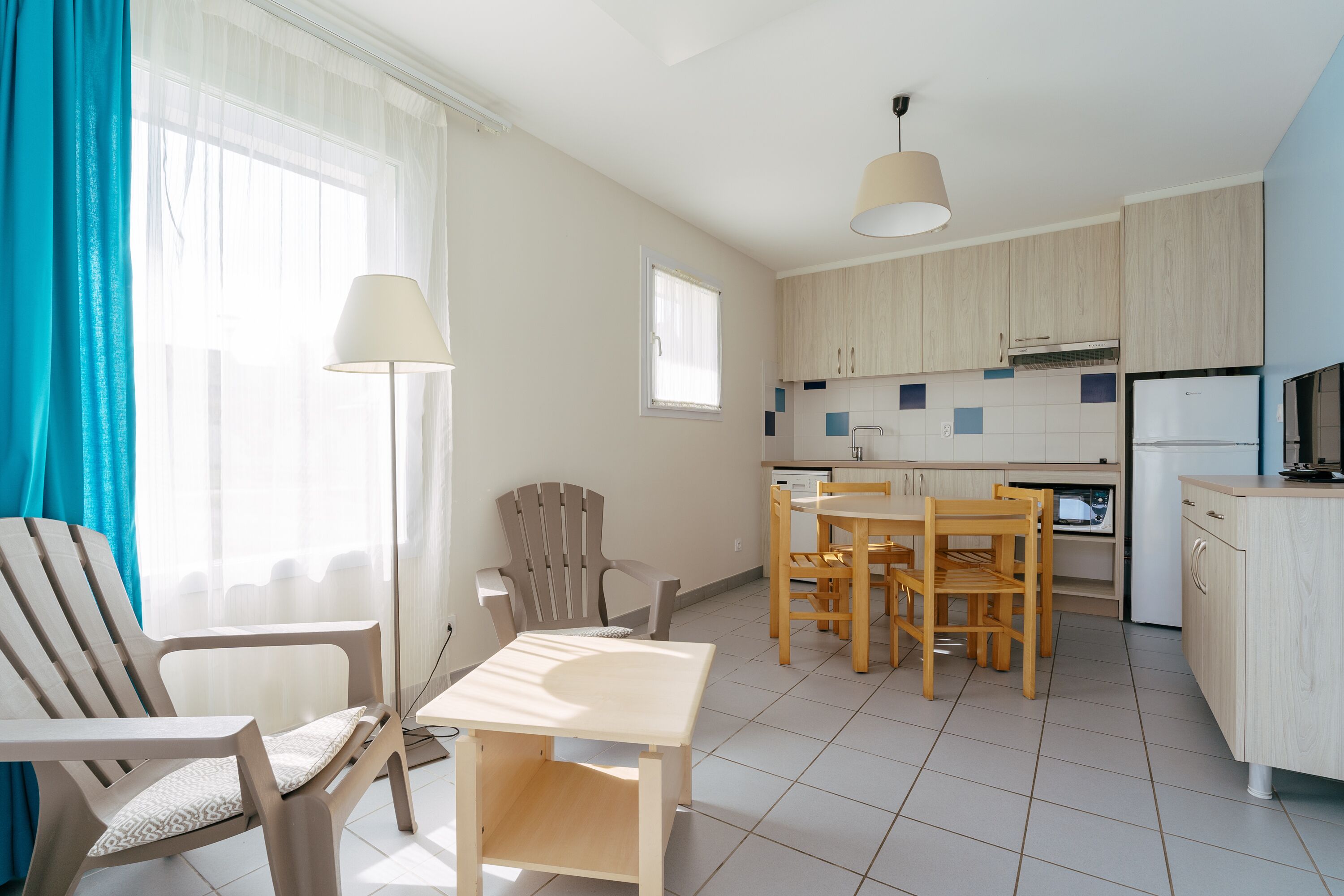 A modern kitchen with wooden cabinets, a dining area with a table and chairs, and a bright window with curtains.