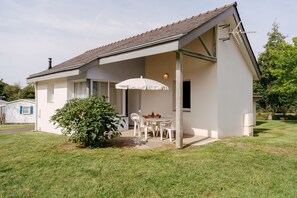 Ein eingeschossiges Haus mit einer überdachten Terrasse, einem Esstisch mit Stühlen und einem kleinen Garten.