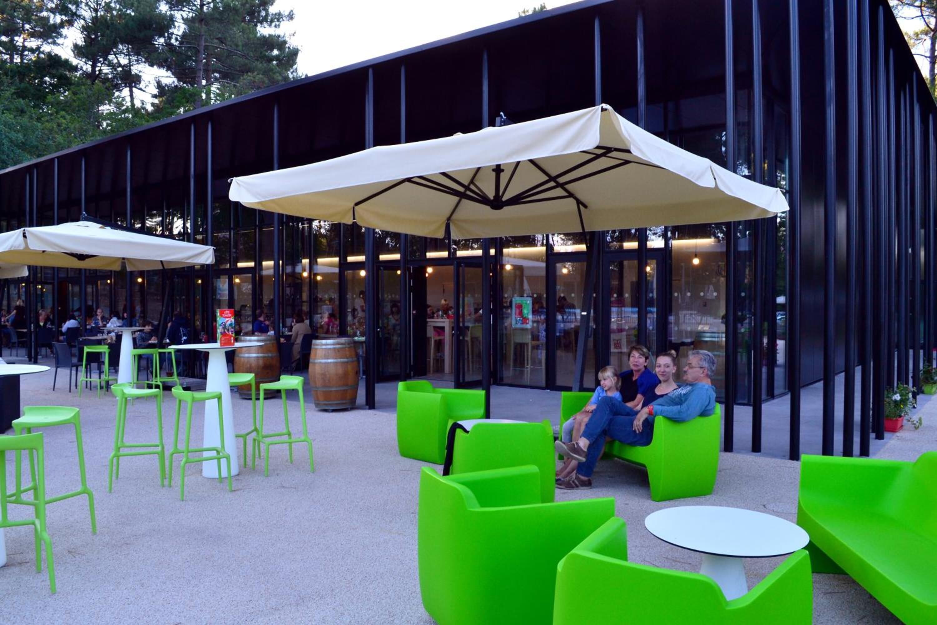 An outdoor seating area with green chairs and tables, covered by large umbrellas, and a modern building with glass walls in the background.
