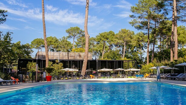 Une piscine à l’eau d’un bleu cristallin, entourée de grands arbres et dotée d’une terrasse en bois avec des chaises longues et des parasols.