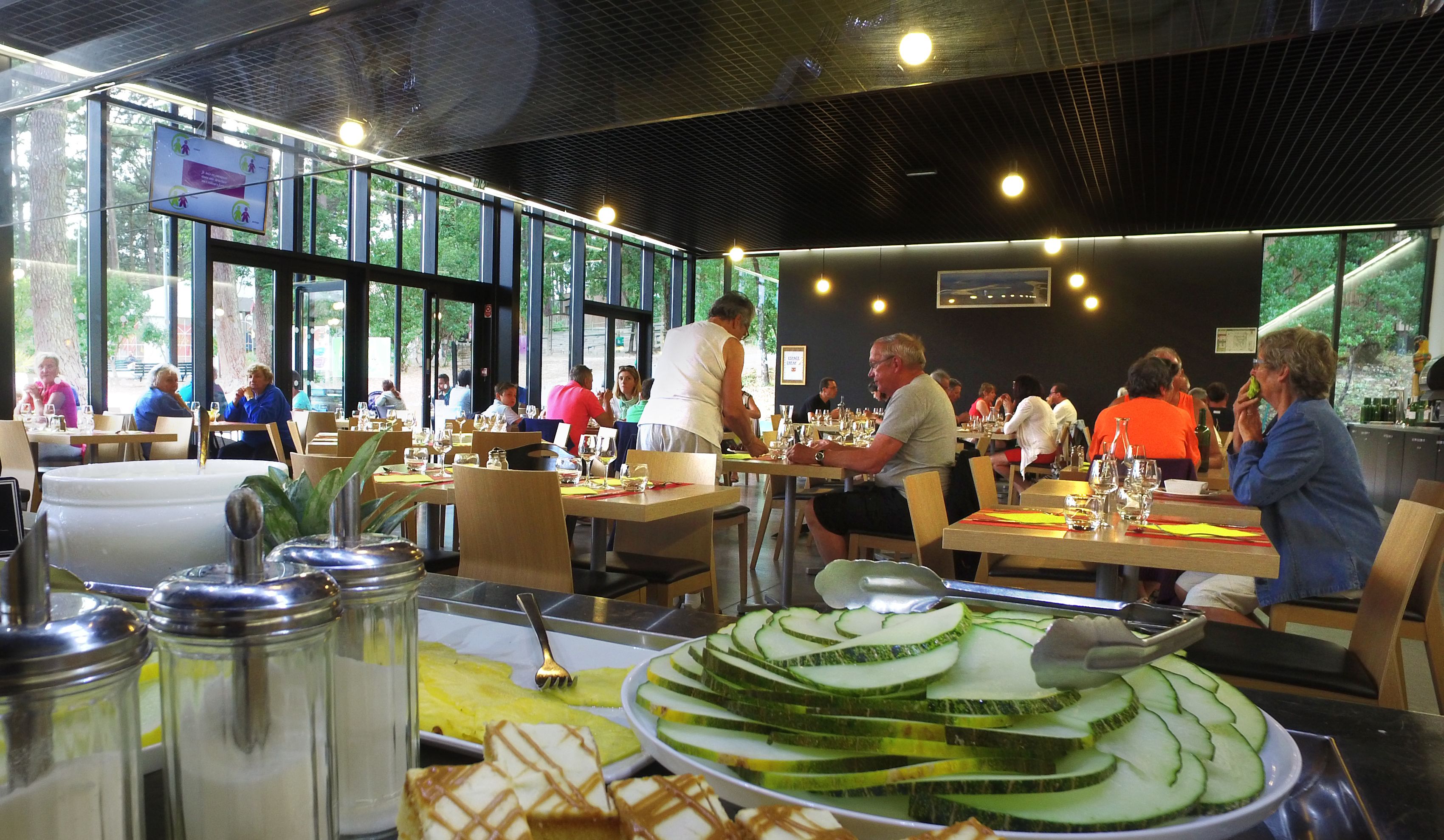 Un restaurante con amplios ventanales, mesas dispuestas con cubiertos y diversos platos, incluyendo pepinos en rodajas y un postre.
