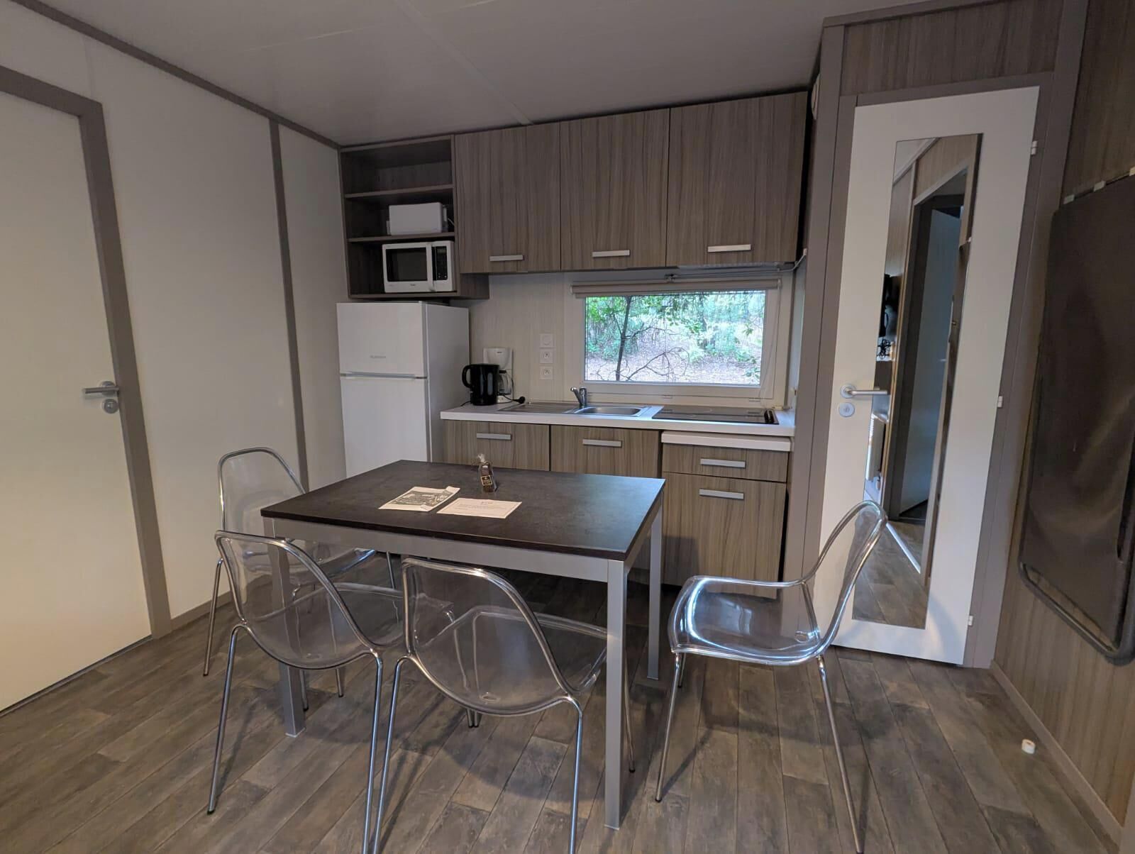 Une petite cuisine équipée d’un four à micro-ondes, d’une machine à café et d’une fenêtre. On y trouve une table avec des chaises et une porte qui donne sur une autre pièce.