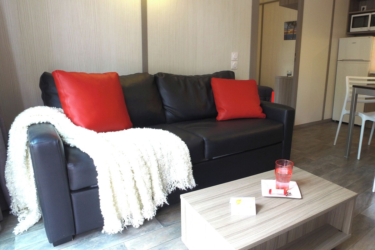 Een woonkamer met een zwarte bank, rode kussens, een witte plaid, een houten salontafel met een glas en een kaart, en een keukenhoek met een magnetron en koelkast.