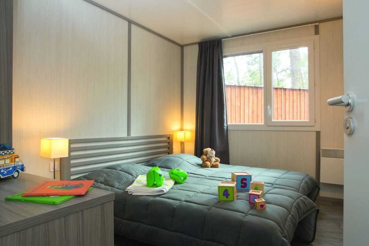 Ein Schlafzimmer mit Bett, Nachttisch, Lampe, Teddybär und einem Fenster mit Vorhängen.