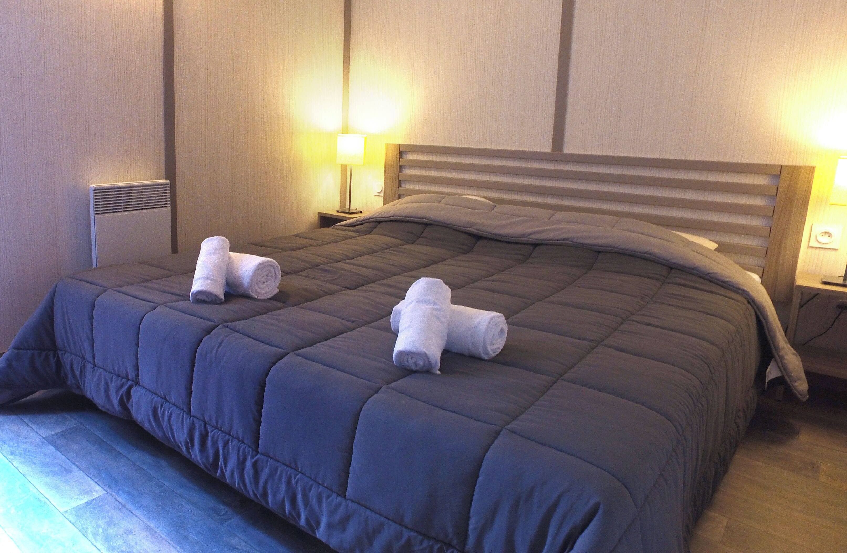A neatly made bed with a dark blue quilt and two rolled towels on the side.