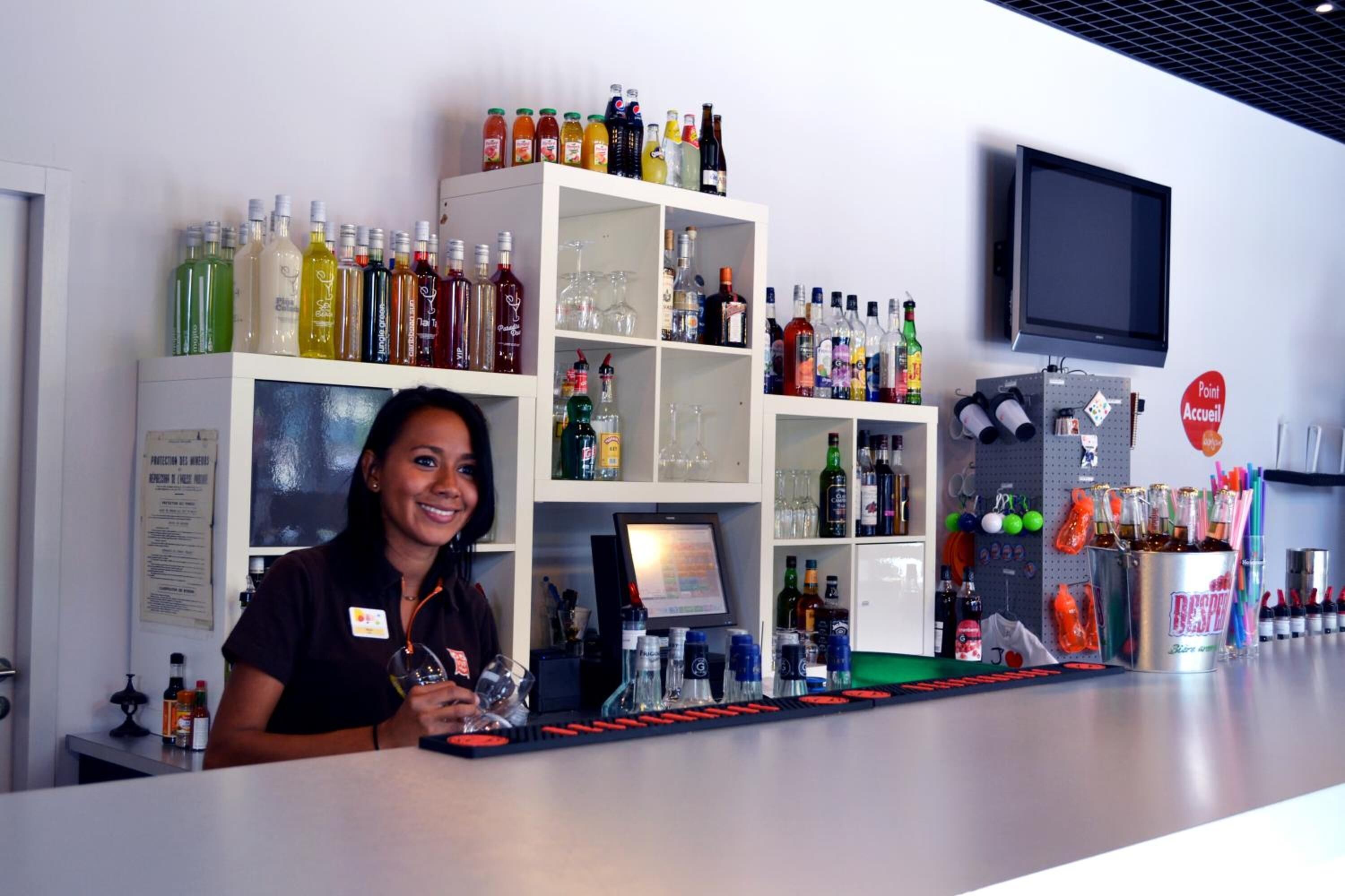 Zona de bar con estantes llenos de botellas de licores, una pantalla digital y un letrero que indica un punto de venta.