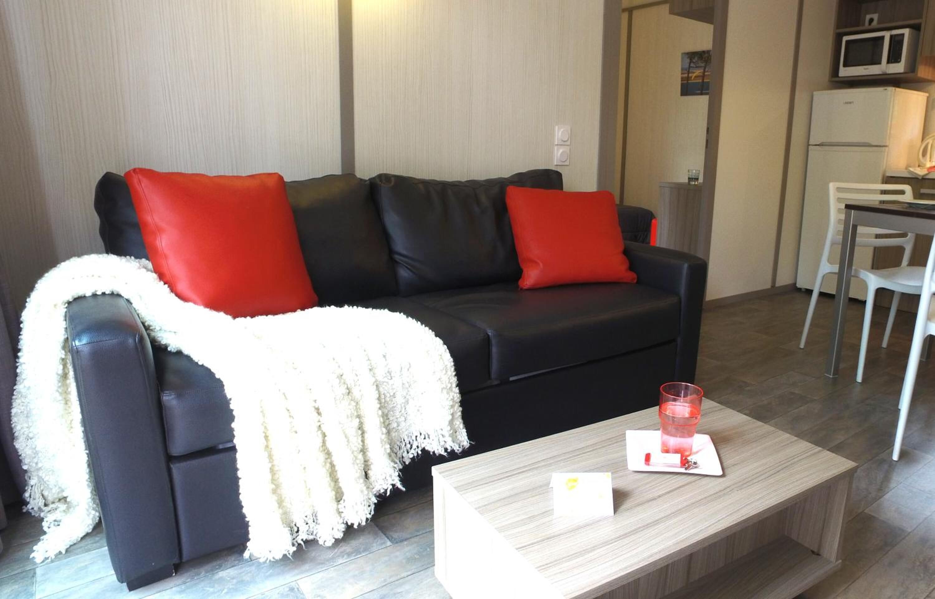 Un salon moderne avec un canapé noir, des coussins rouges, un plaid blanc et une table basse en bois sur laquelle se trouvent un verre et une carte.