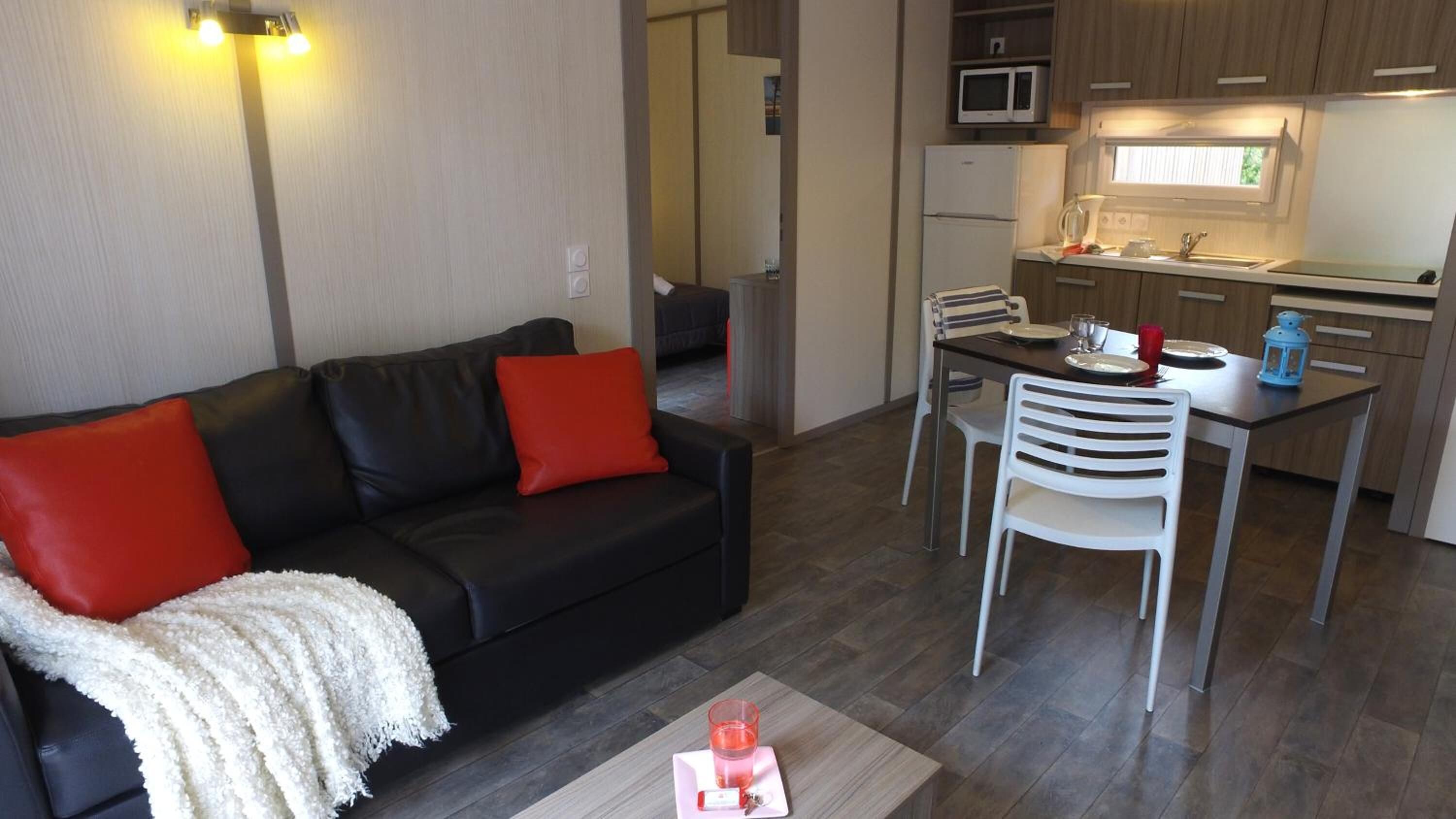 Un espacio habitable compacto con un sofá negro, cojines rojos, una manta blanca, una mesa de comedor con sillas, un microondas, un refrigerador y una ventana con persianas.