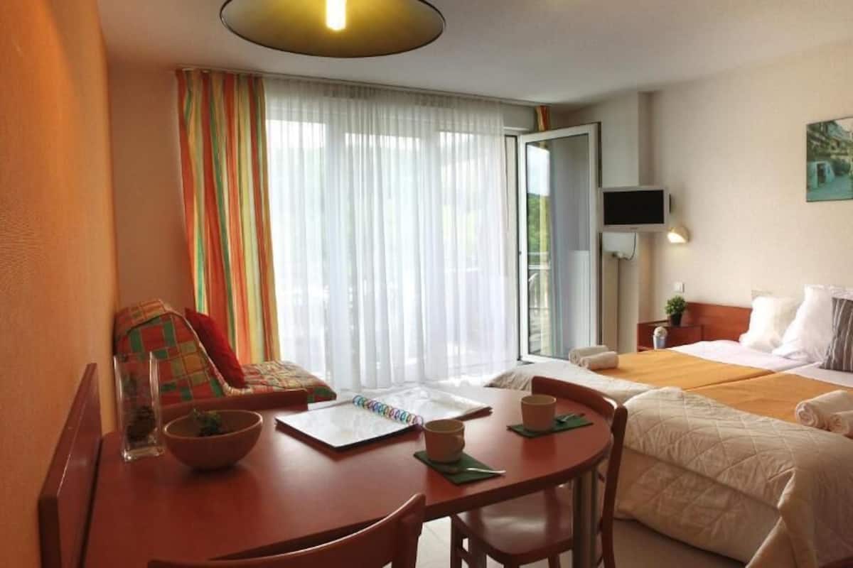 A hotel room with a bed, a desk with a chair, a table, a bowl, a plant, and a window with curtains.