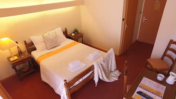A neatly made bed with white linens, a bedside table with a lamp, a small potted plant, and a wooden chair.