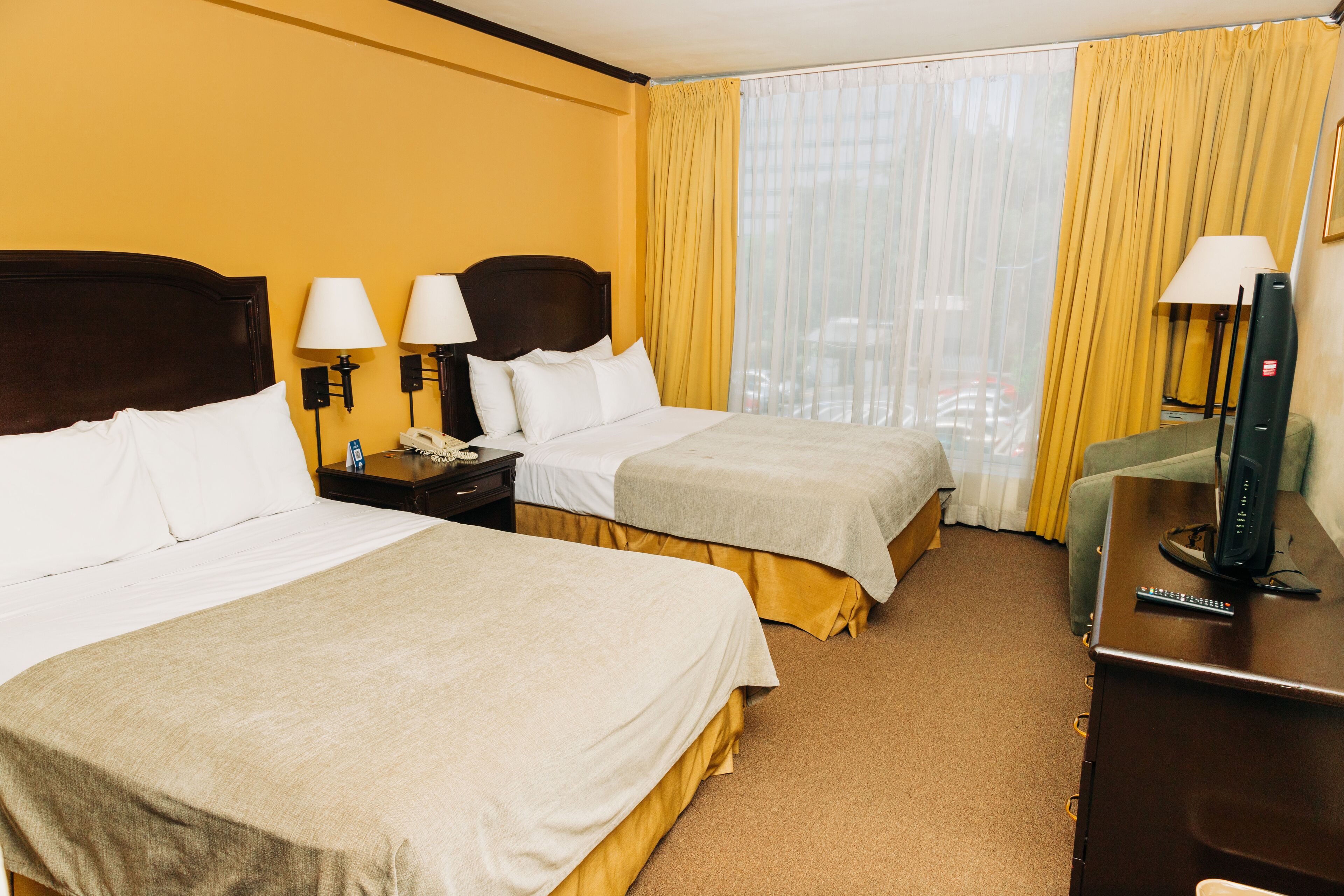 Une chambre d’hôtel avec deux lits, un bureau avec une télévision et une fenêtre avec des rideaux jaunes.