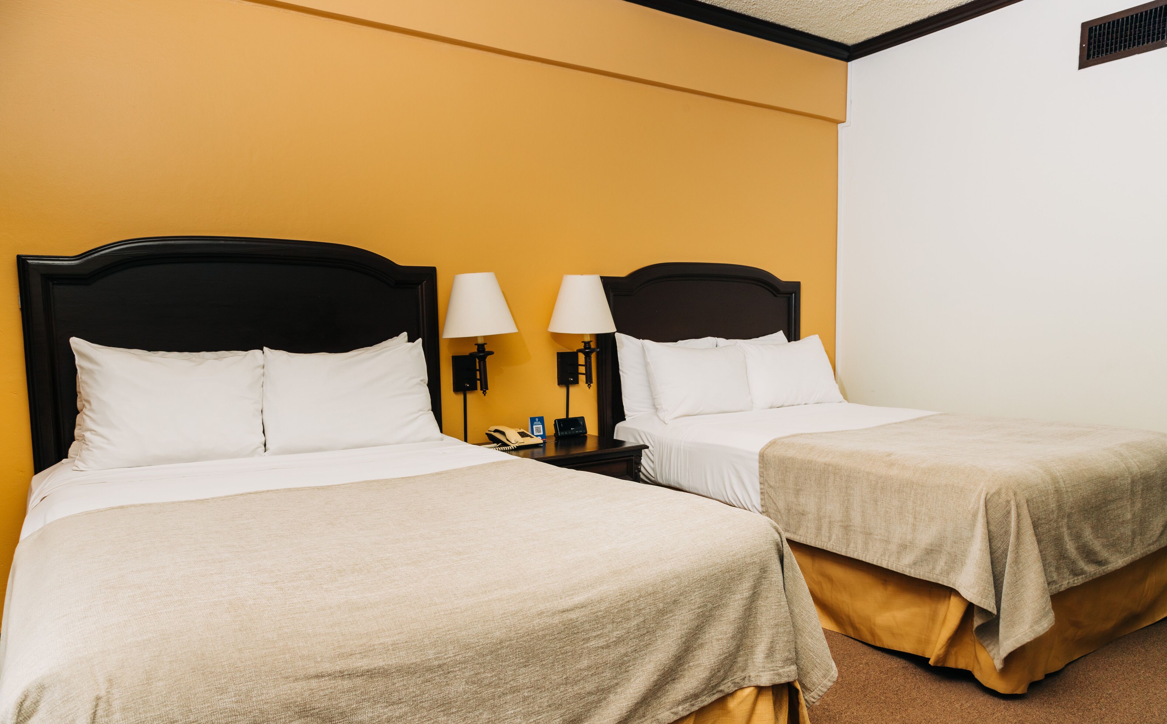 Une chambre d’hôtel avec deux lits, chacun étant équipé de draps blancs et d’une couverture beige. Il y a deux tables de chevet avec des lampes et un téléphone. Les murs sont de couleur jaune et la chambre présente un aménagement simple et épuré.