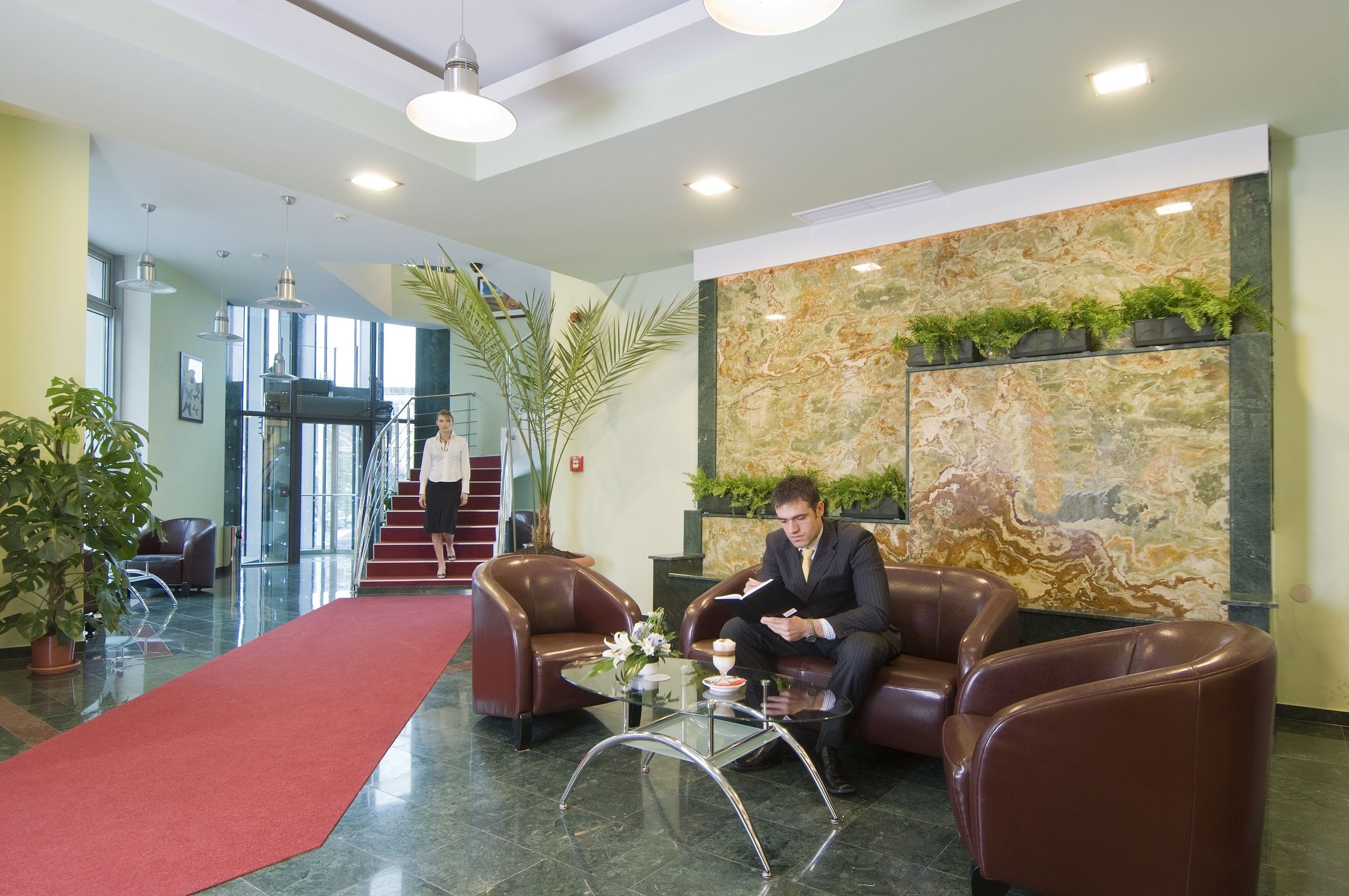 A lobby with leather chairs, a red carpet, and a marble wall with plants.