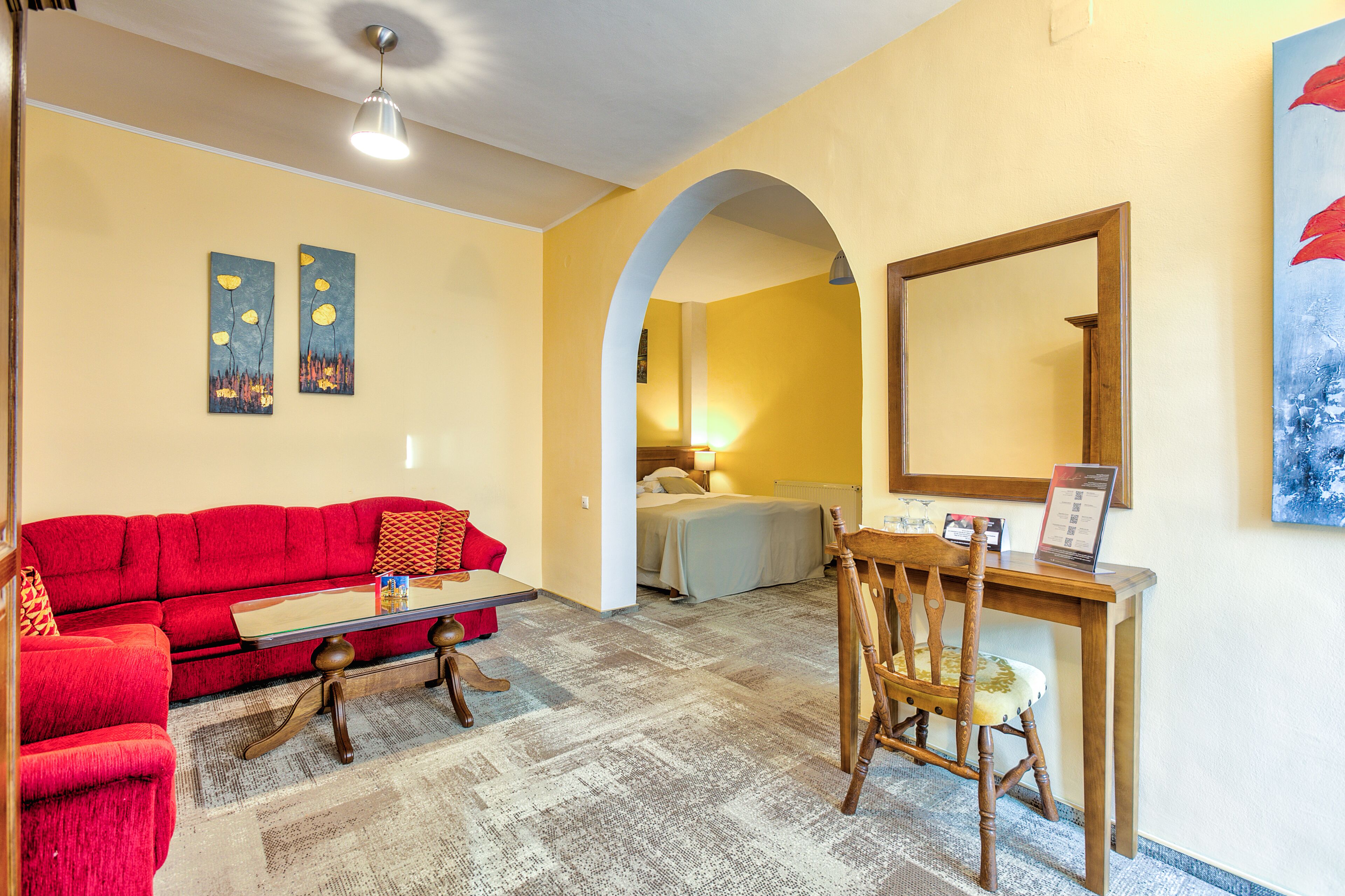 A hotel room with a red sofa, a wooden desk, a chair, a bed, and two paintings on the wall.