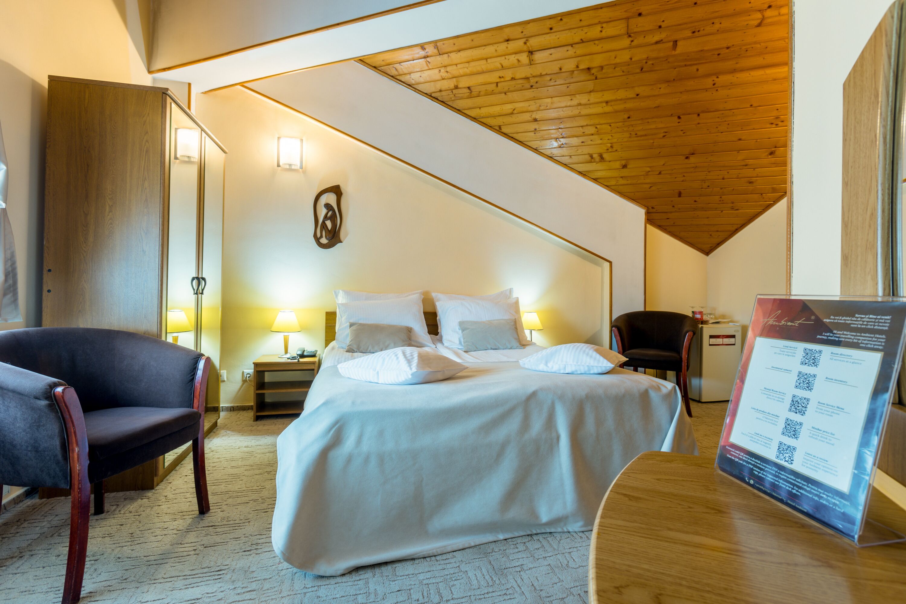 A hotel room with a bed, a chair, a desk, and a wooden ceiling.