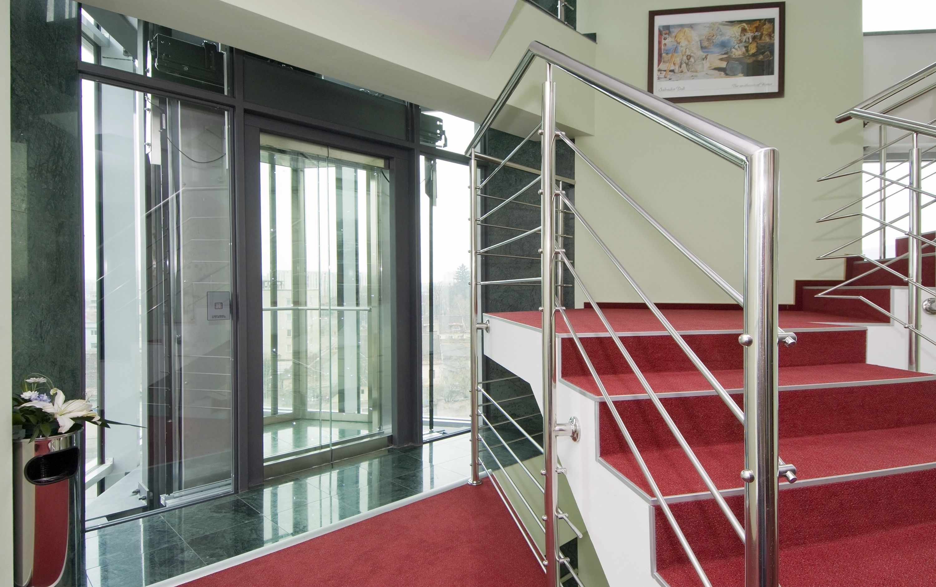 A modern interior with a glass elevator, red carpeted stairs, and a framed artwork on the wall.