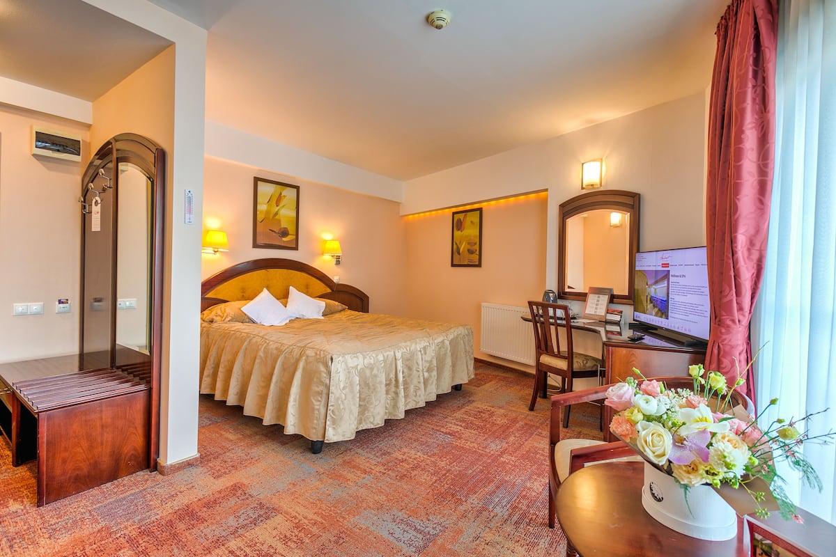 A hotel room with a bed, a desk with a computer, a chair, a mirror, and a window with curtains.