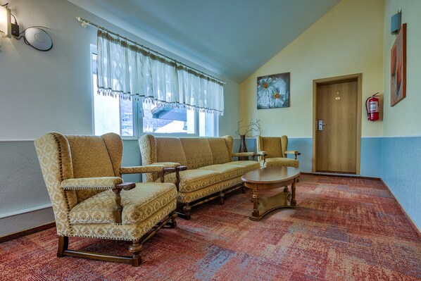 A room with a patterned carpet, two armchairs, a sofa, a small table, a wooden door, and a window with sheer curtains.