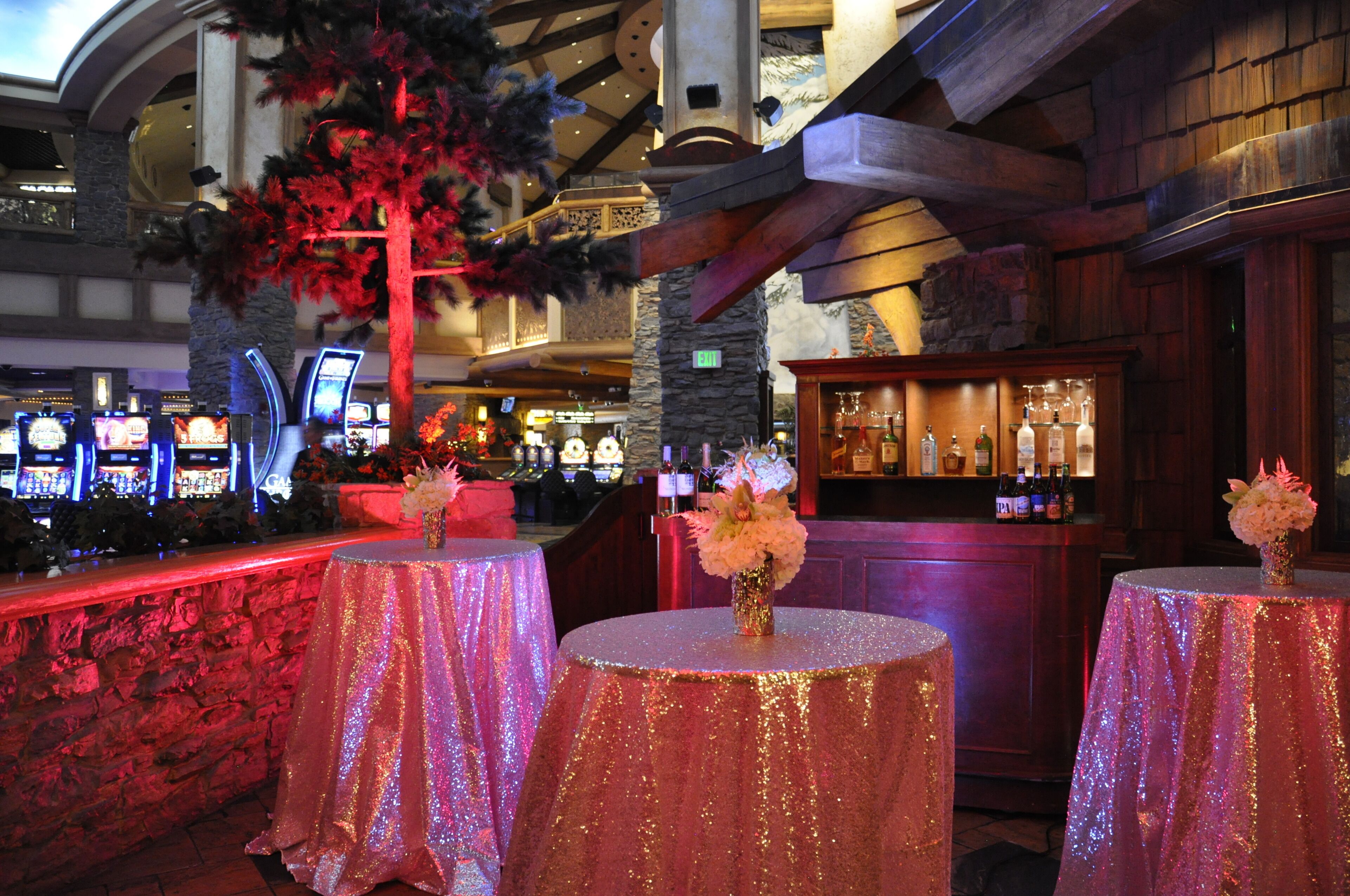 A bar area with round tables covered in glittery pink tablecloths, a wooden bar counter, and a decorative tree with red foliage.