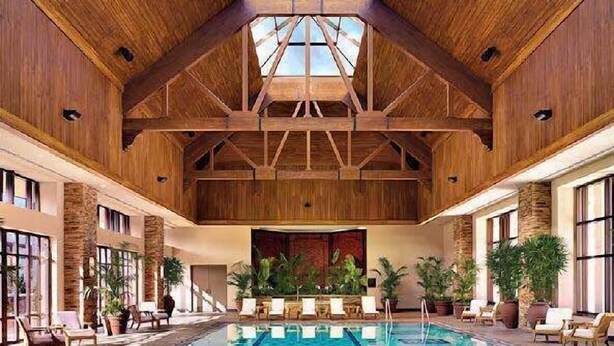 An indoor swimming pool with wooden beams and a high ceiling, surrounded by lounge chairs and potted plants.