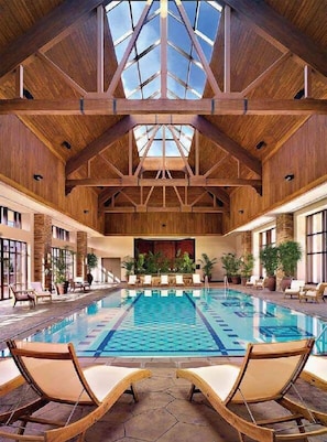 Una piscina interior con vigas de madera y techo alto, rodeada de sillones de descanso y plantas en macetas.