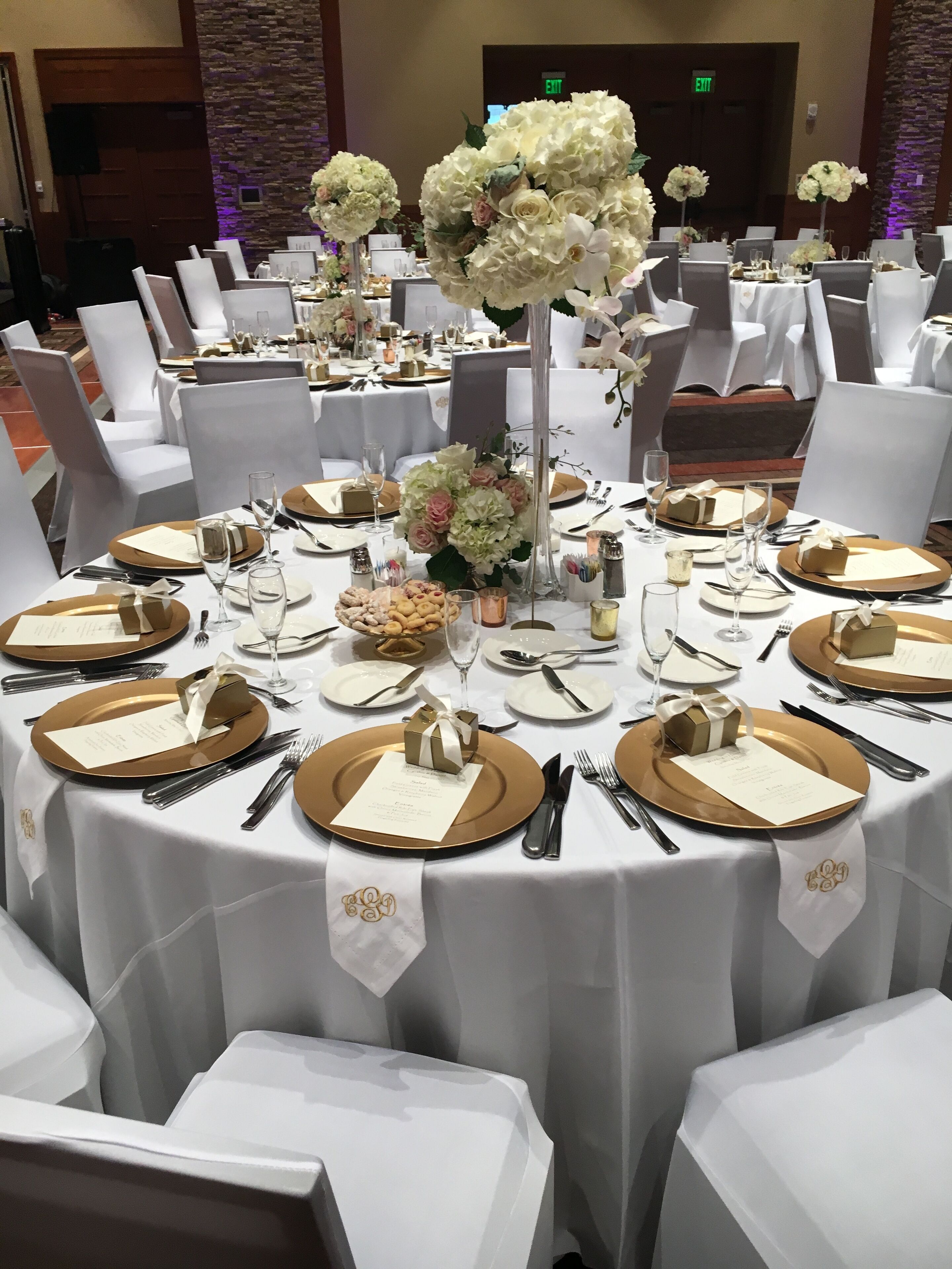 Un salón de banquetes con mesas redondas dispuestas para un evento formal, con manteles blancos, platos bajos dorados y centros de mesa florales.
