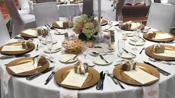 Un salón de banquetes con mesas redondas dispuestas para un evento formal, con manteles blancos, platos bajos dorados y centros de mesa florales.