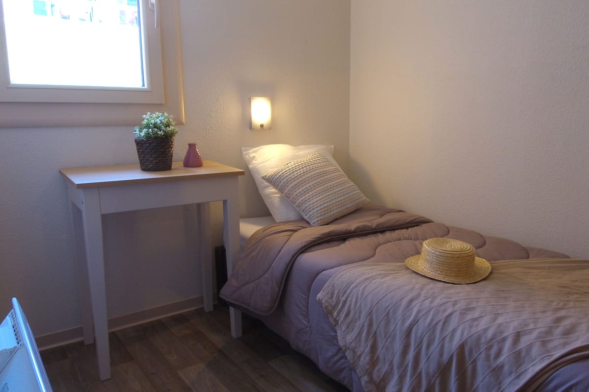 A small, neatly arranged bedroom with a single bed, a bedside table, a lamp, and a window.