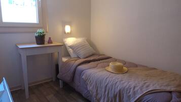 A small, neatly arranged bedroom with a single bed, a bedside table, a lamp, and a window.