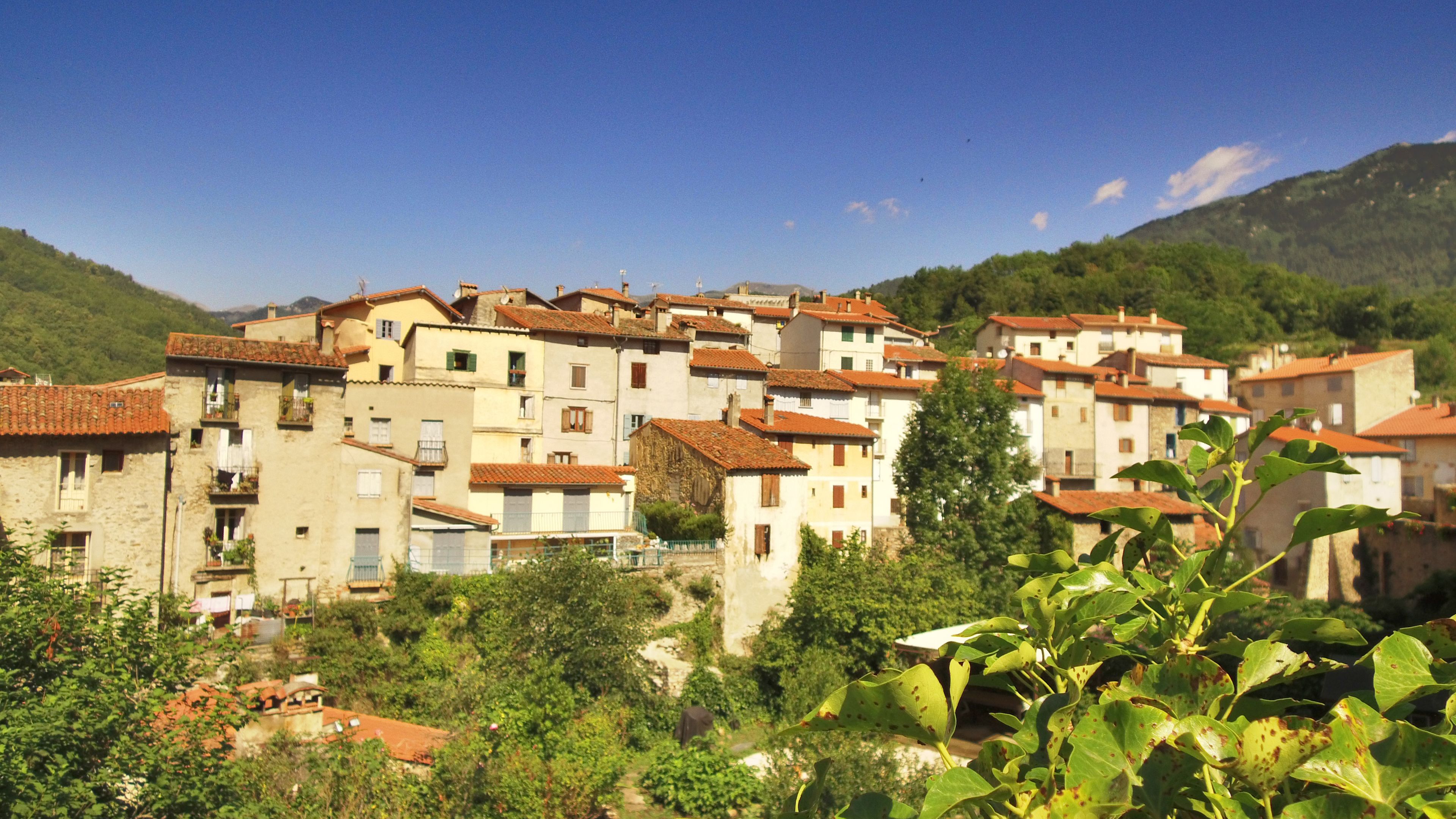 A hillside village with terracotta-roofed buildings, lush greenery, and a clear blue sky.