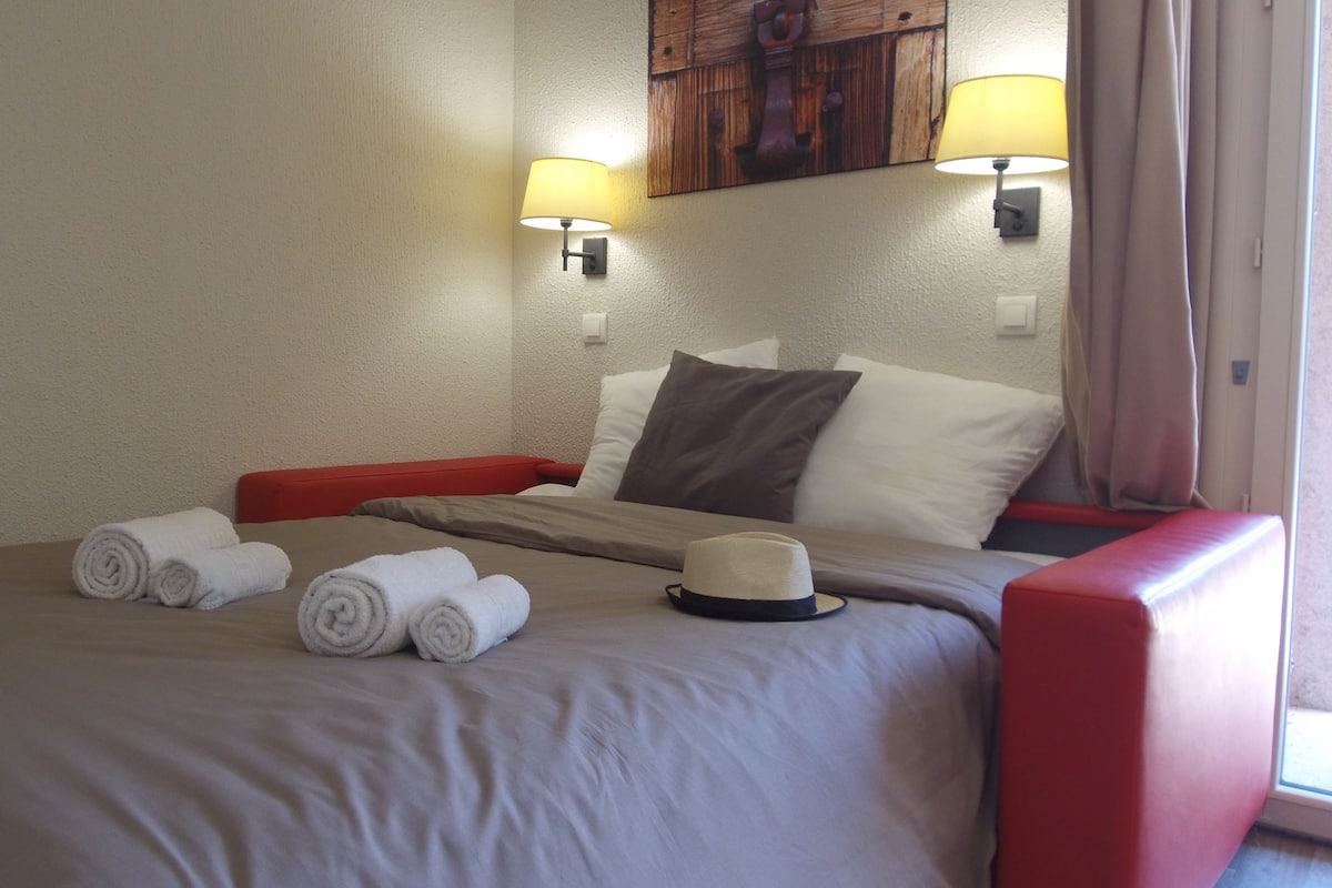 A hotel room with a bed, two wall-mounted lamps, a red armchair, and a window with curtains.