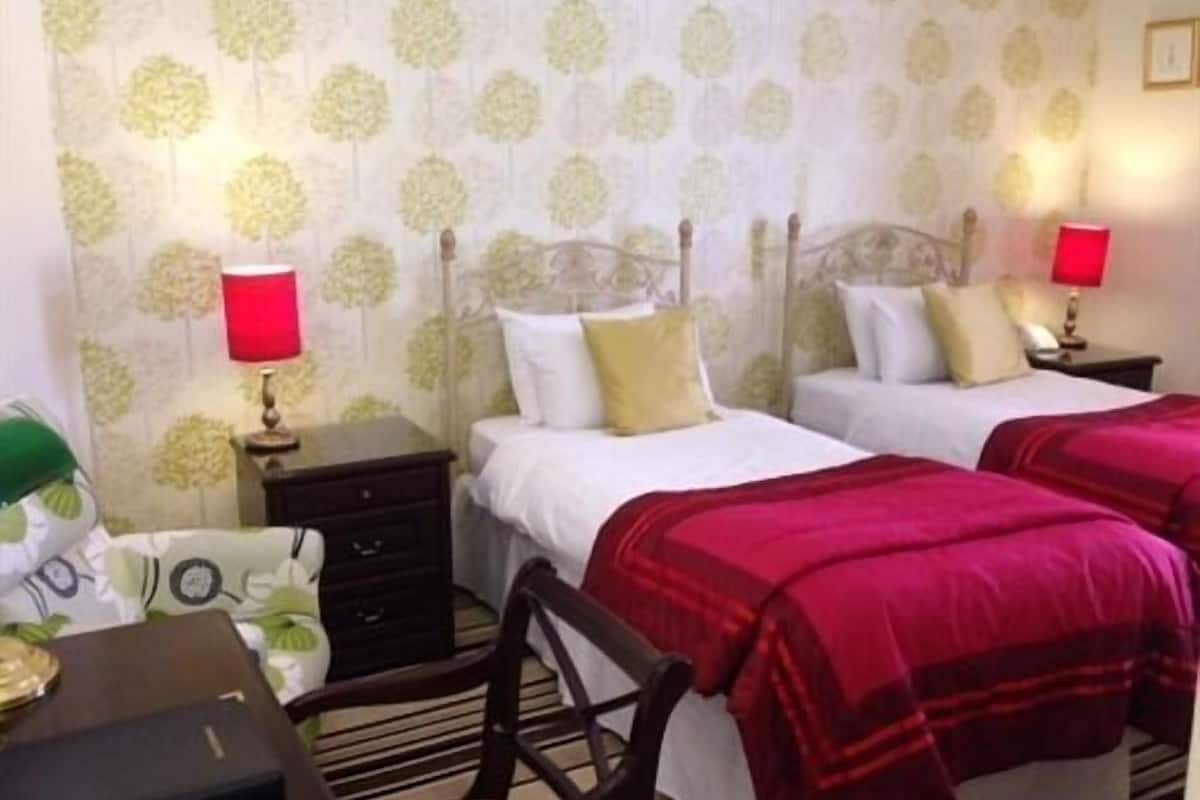 A hotel room with two beds, each with white linens and red blankets. There are two bedside tables with lamps, a chair, and a desk with a lamp. The walls have a floral wallpaper pattern.