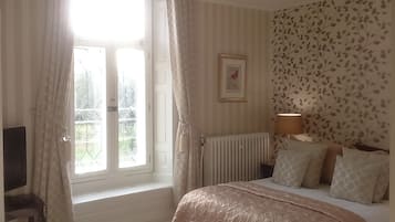 A bedroom with a bed, a radiator, a window with curtains, a wall with a leaf pattern, and a framed picture.