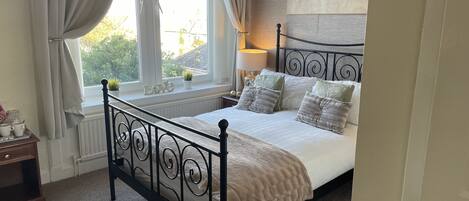 Un dormitorio con cama, una ventana con cortinas, una mesita de noche y un cuadro floral en la pared.