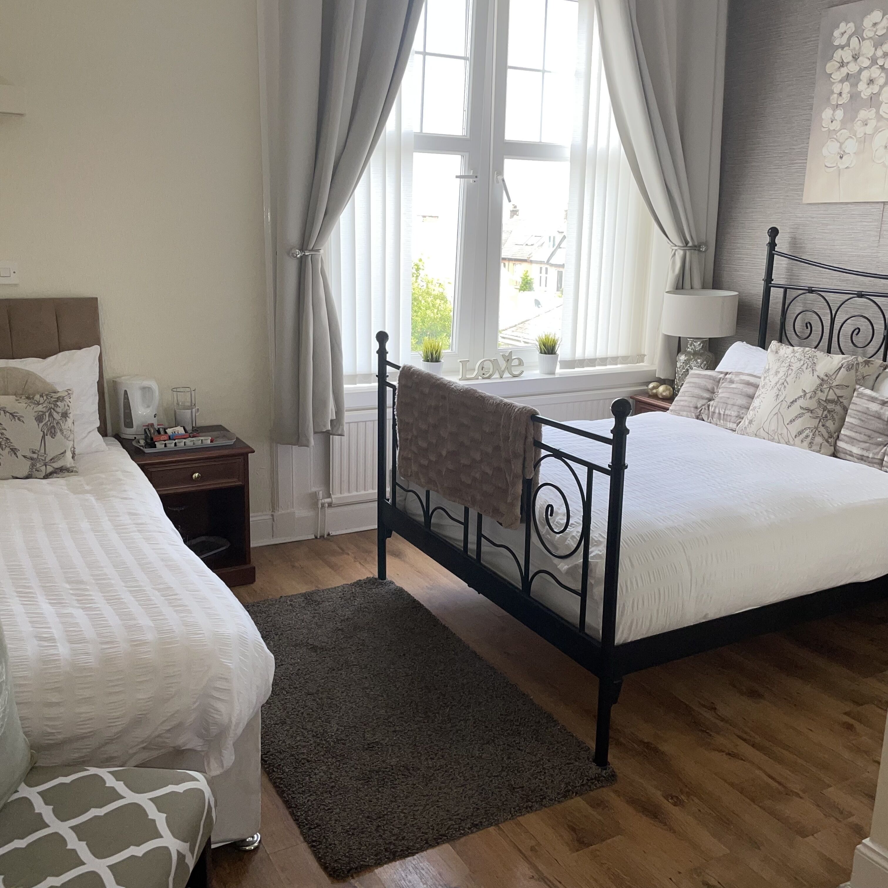 A bedroom with two beds, a window with curtains, a bedside table with a lamp, and a view of the outside.