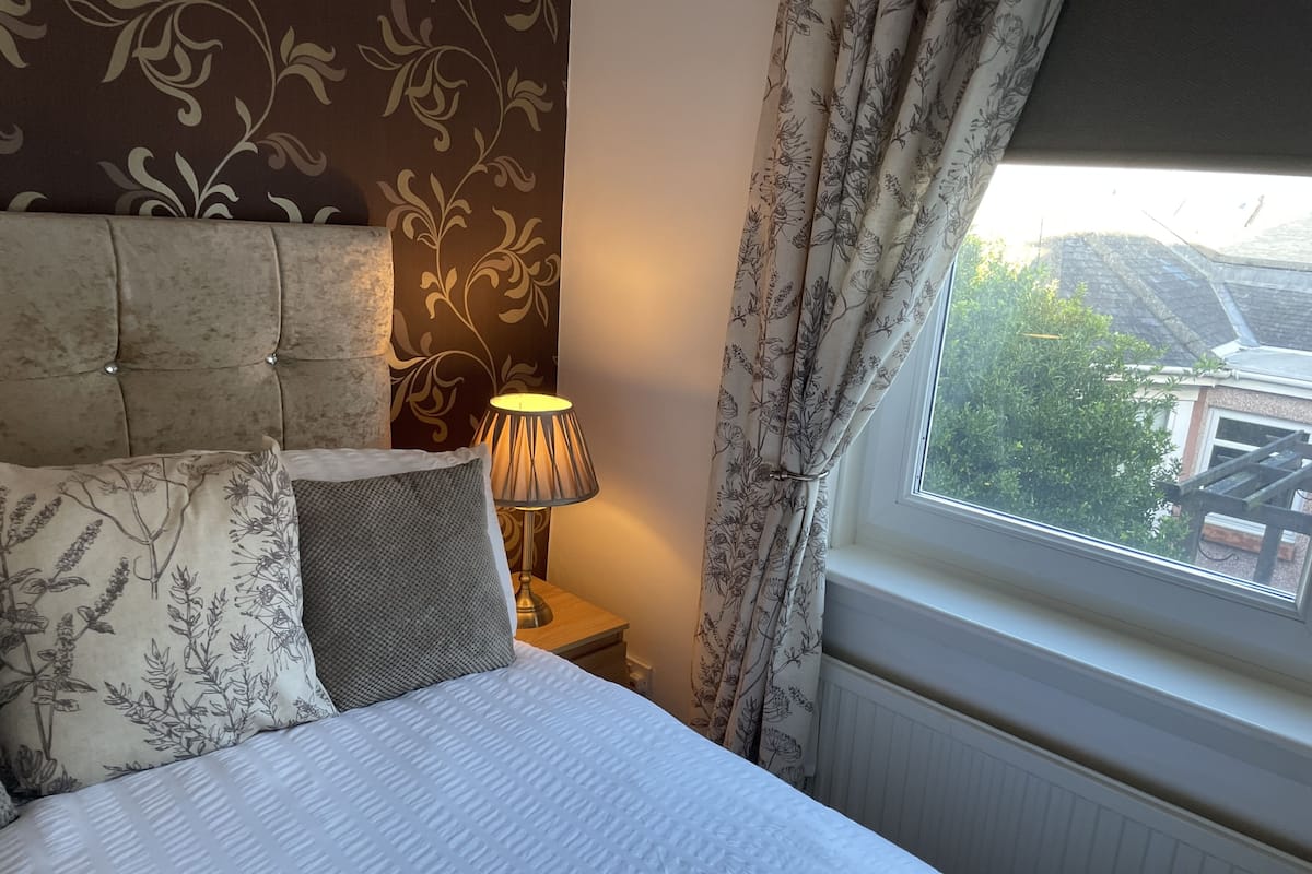 A neatly made bed with floral-patterned pillows, a bedside lamp, and a view of greenery outside a window.