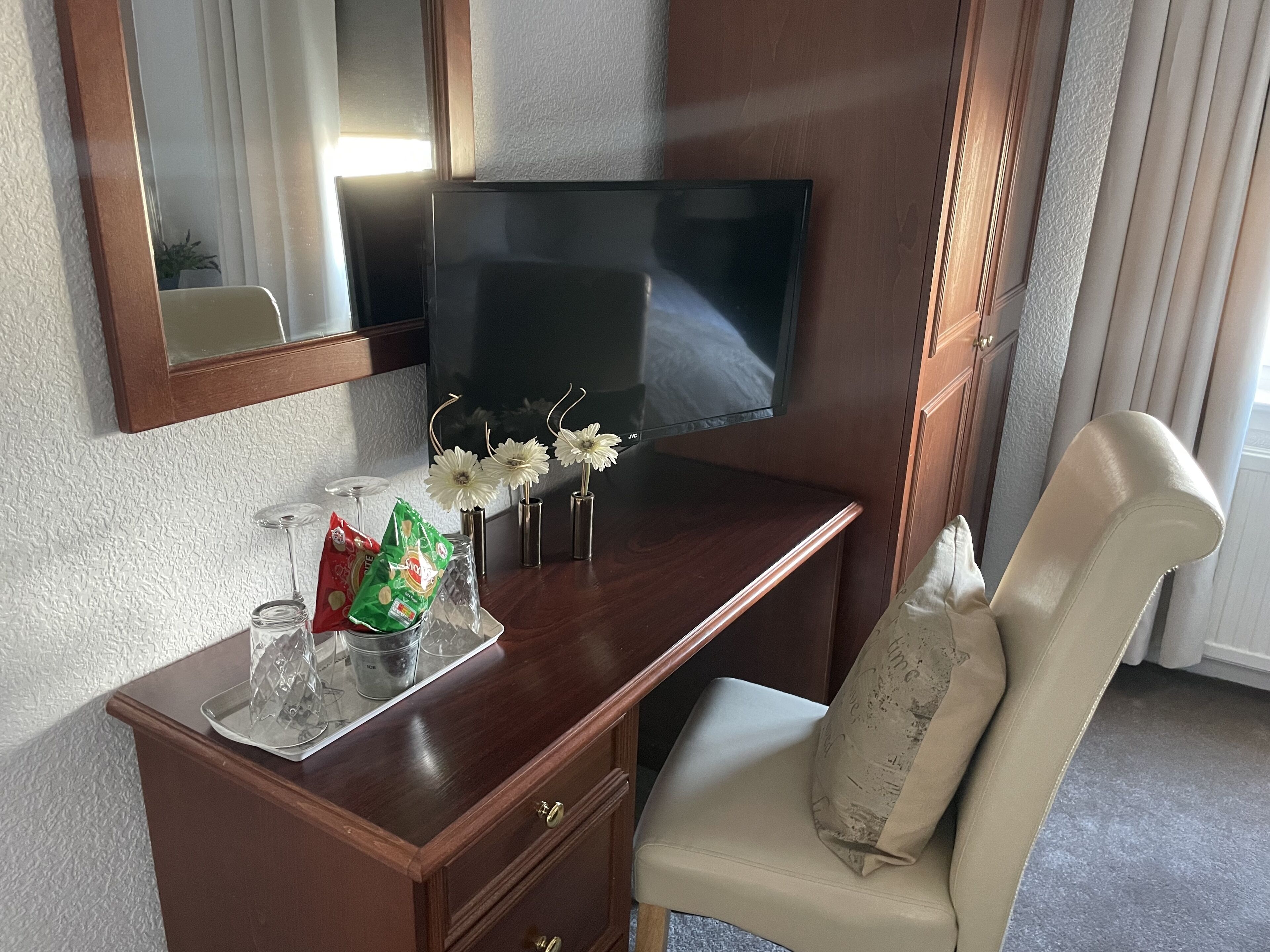 A hotel room with a wooden desk, a flat-screen TV, a mirror, a vase with white flowers, a glass of water, and a chair with a cushion.