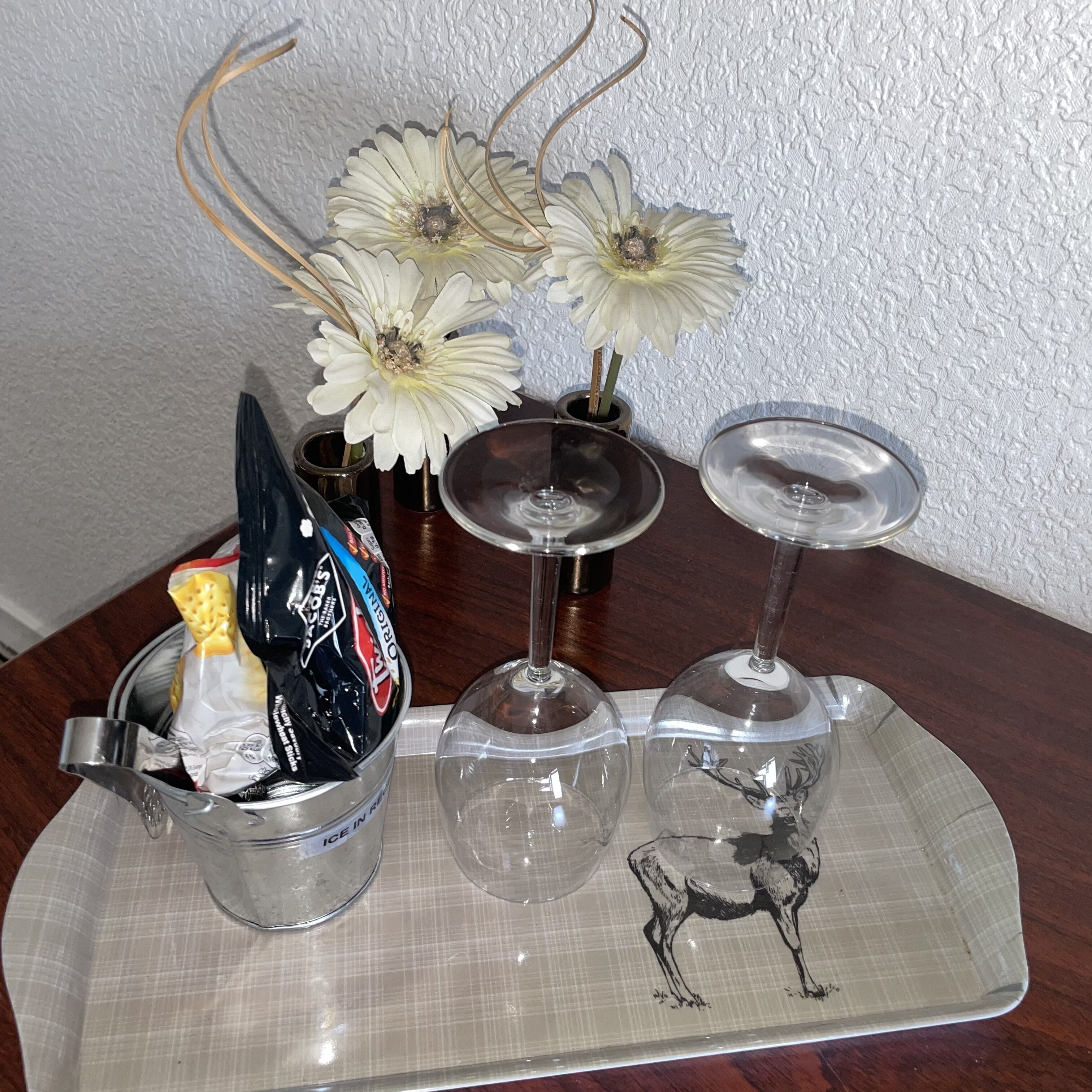 Un vassoio con tre bicchieri da vino, un piccolo vaso con fiori bianchi e un contenitore con snack.