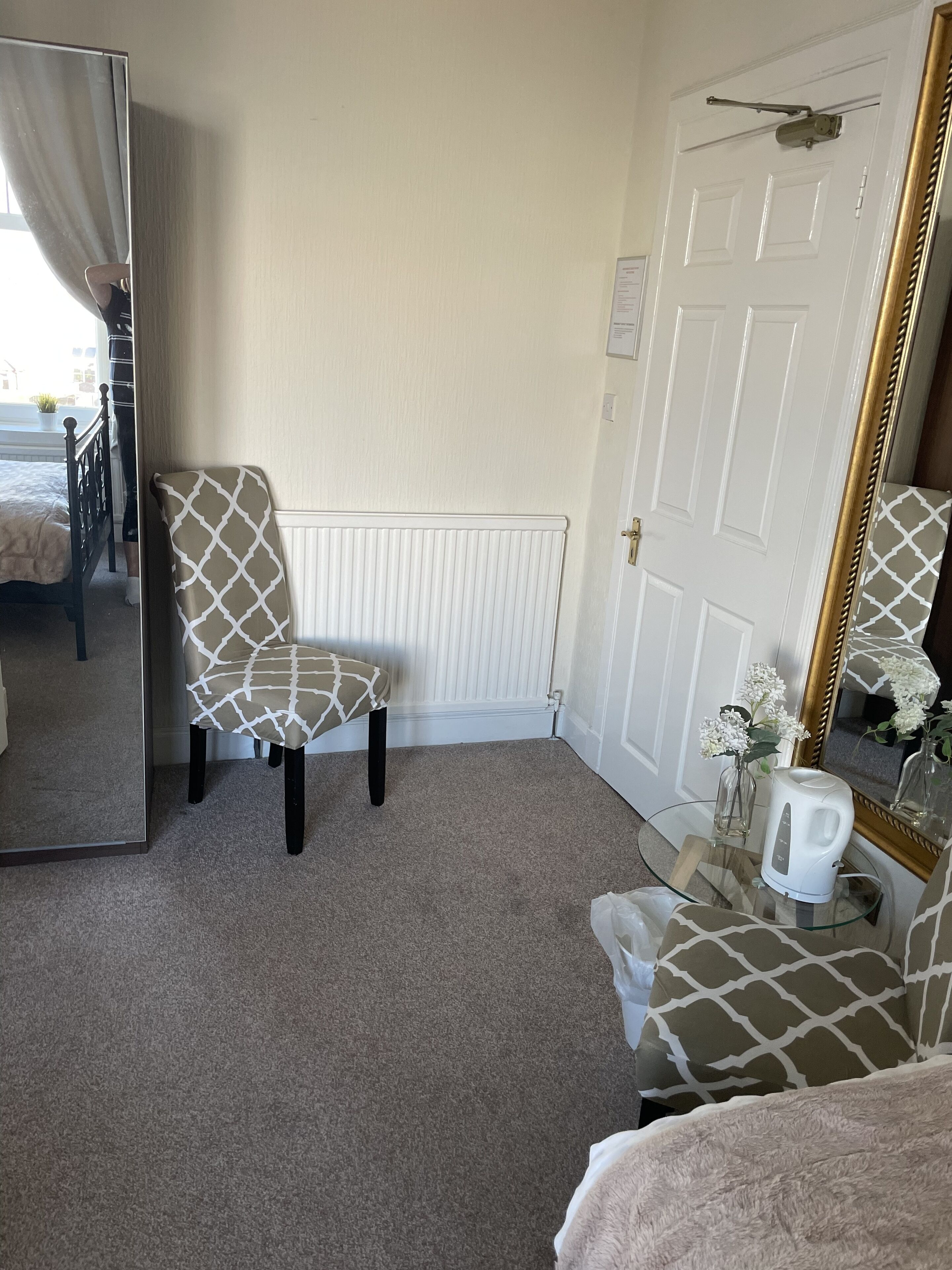 A bedroom with a bed, a chair, a mirror, a radiator, and a small table with a kettle.
