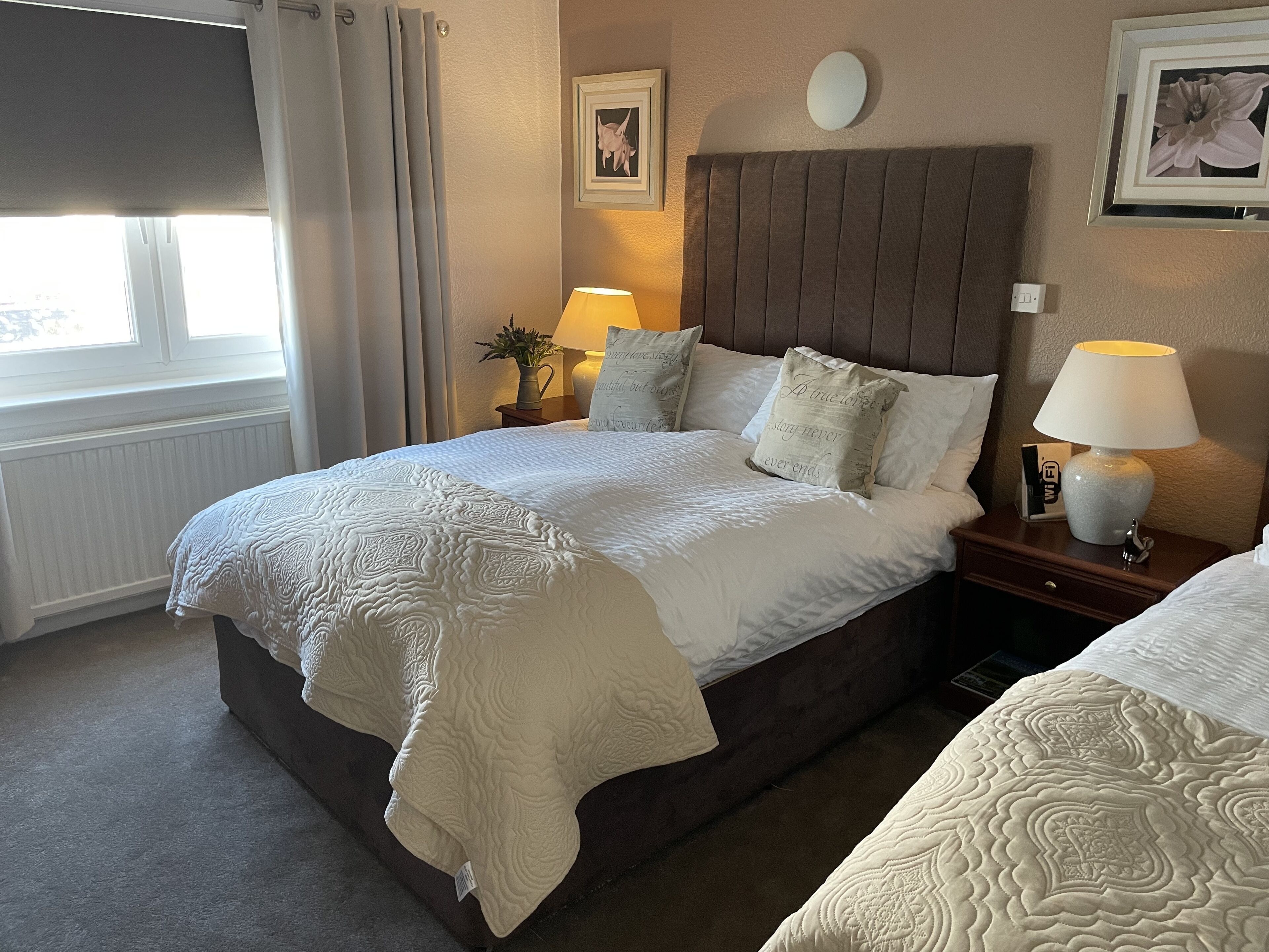 A neatly made bed with a headboard, two bedside lamps, a nightstand, and a window with curtains.