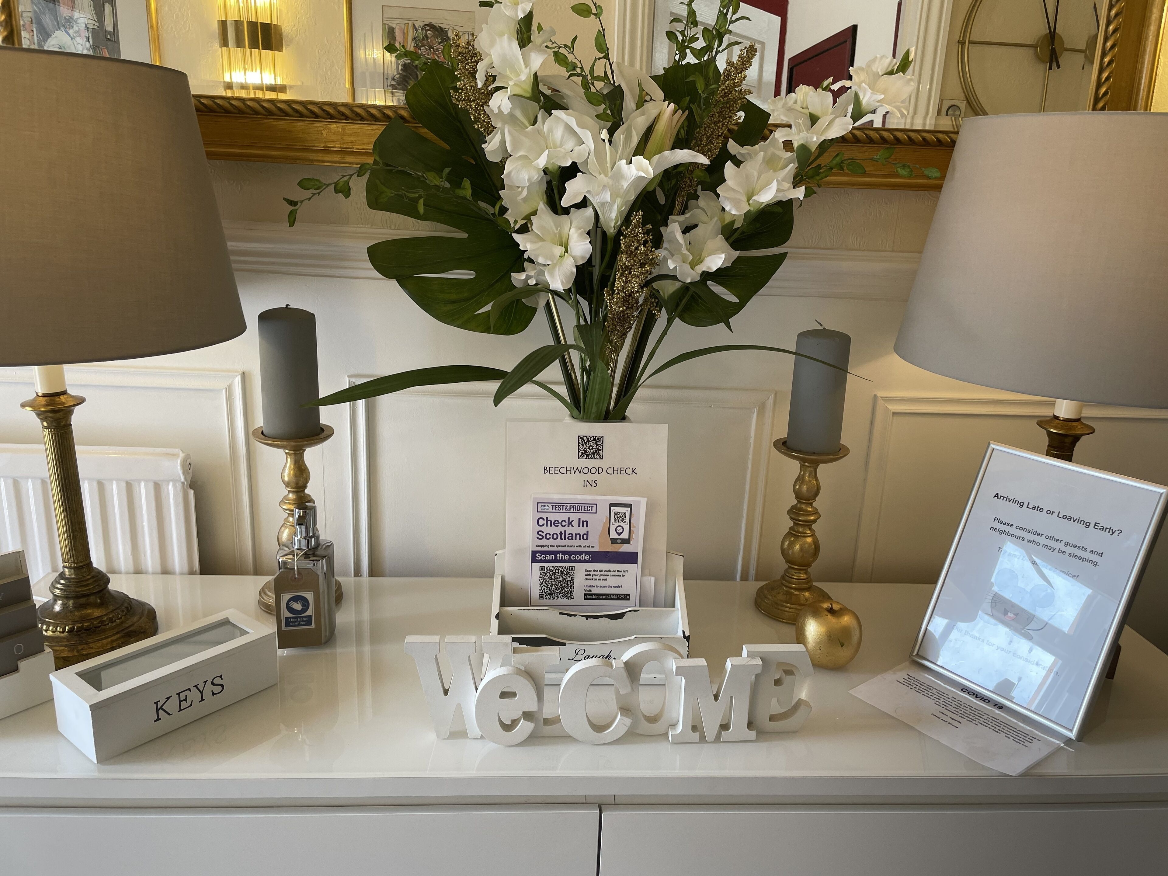 A reception desk with a 'Welcome' sign, a 'Keys' sign, a QR code for check-in, and a laptop displaying COVID-19 information.
