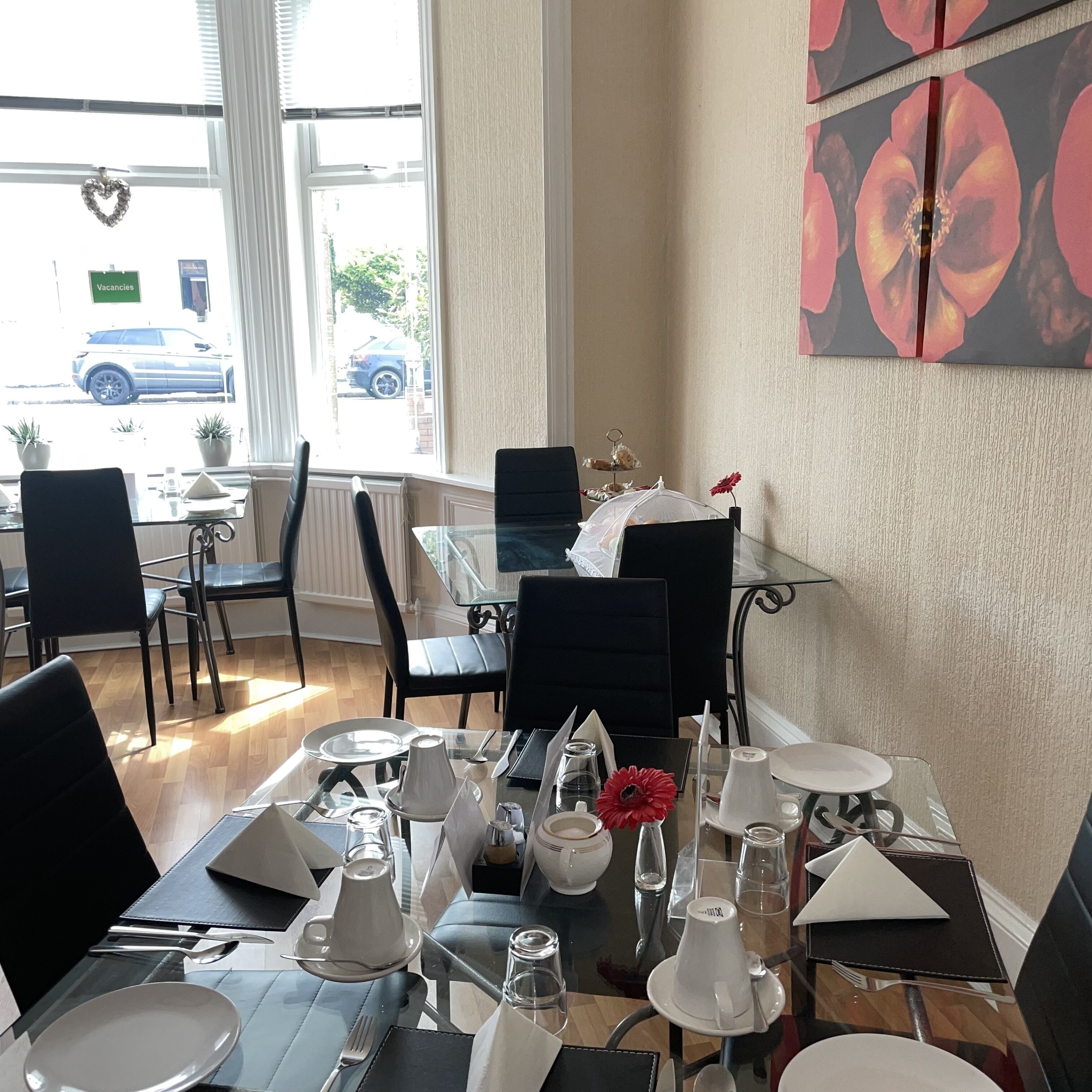 Un coin repas avec une table dressée pour deux personnes, une fenêtre donnant sur la rue et un mur orné d’un tableau représentant des fleurs.