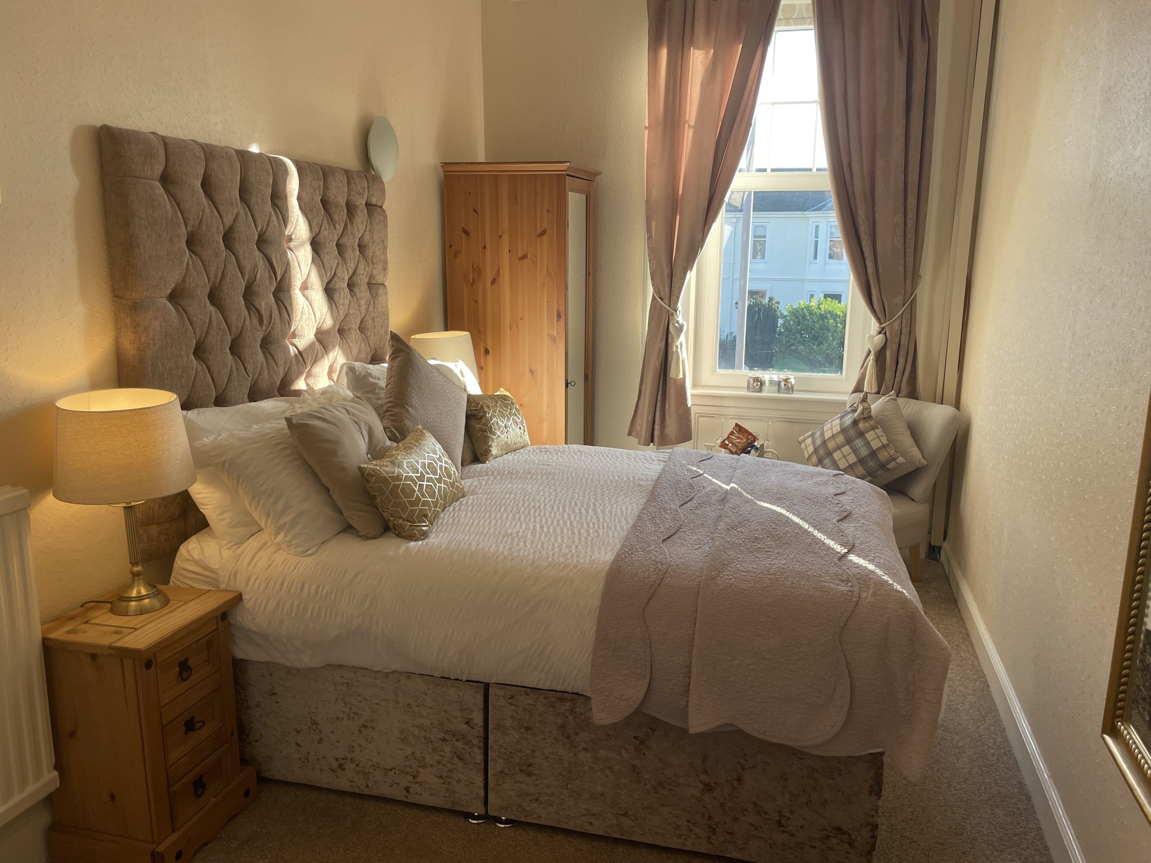 A bedroom with a large bed, a wooden nightstand, a lamp, and a view of the outside through a window with curtains.