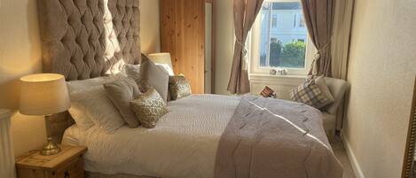A bedroom with a large bed, a wooden nightstand, a lamp, and a view of the outside through a window with curtains.