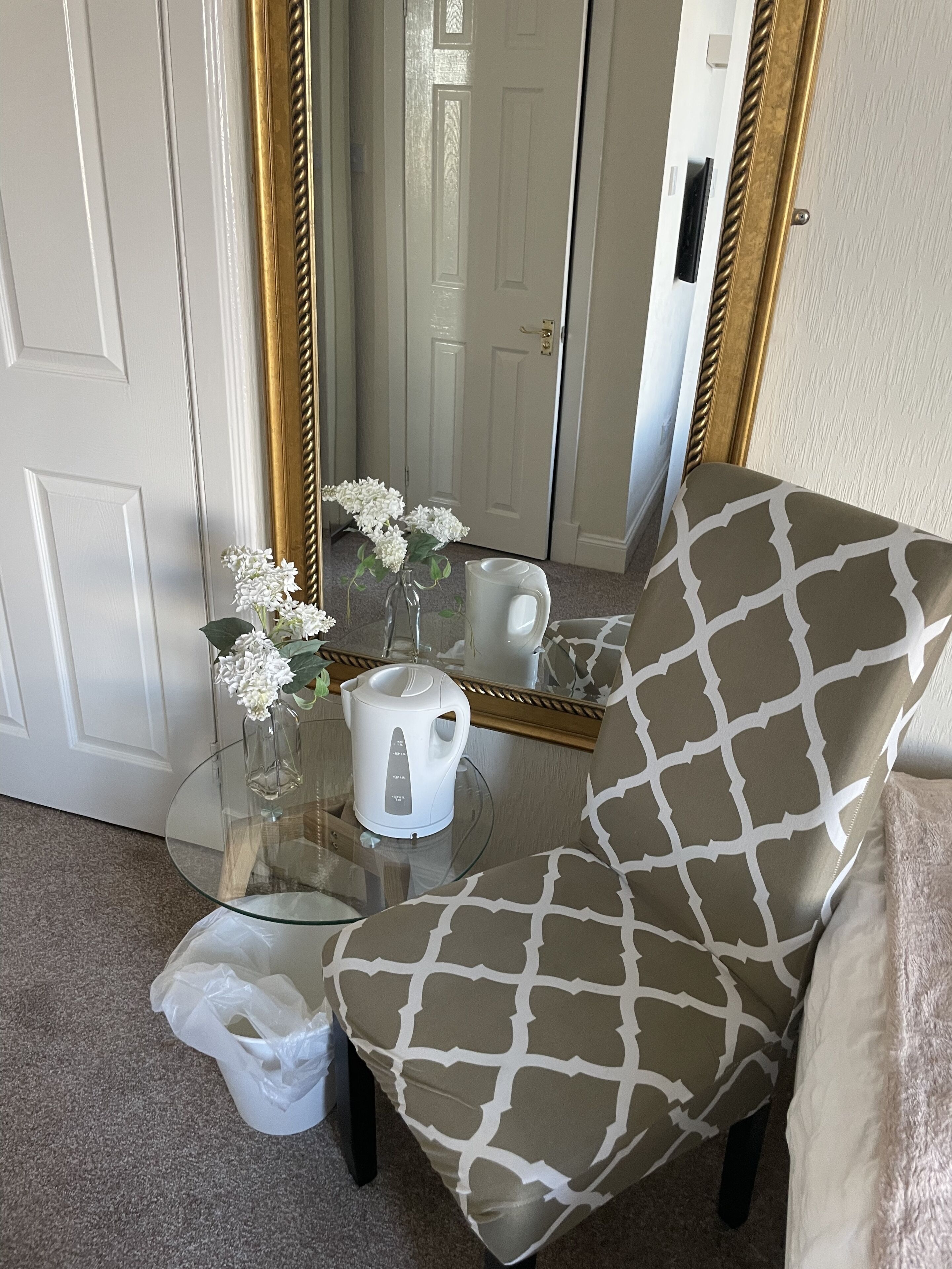 A room with a glass table, a white kettle, a vase with flowers, a patterned armchair, and a mirror reflecting a door.