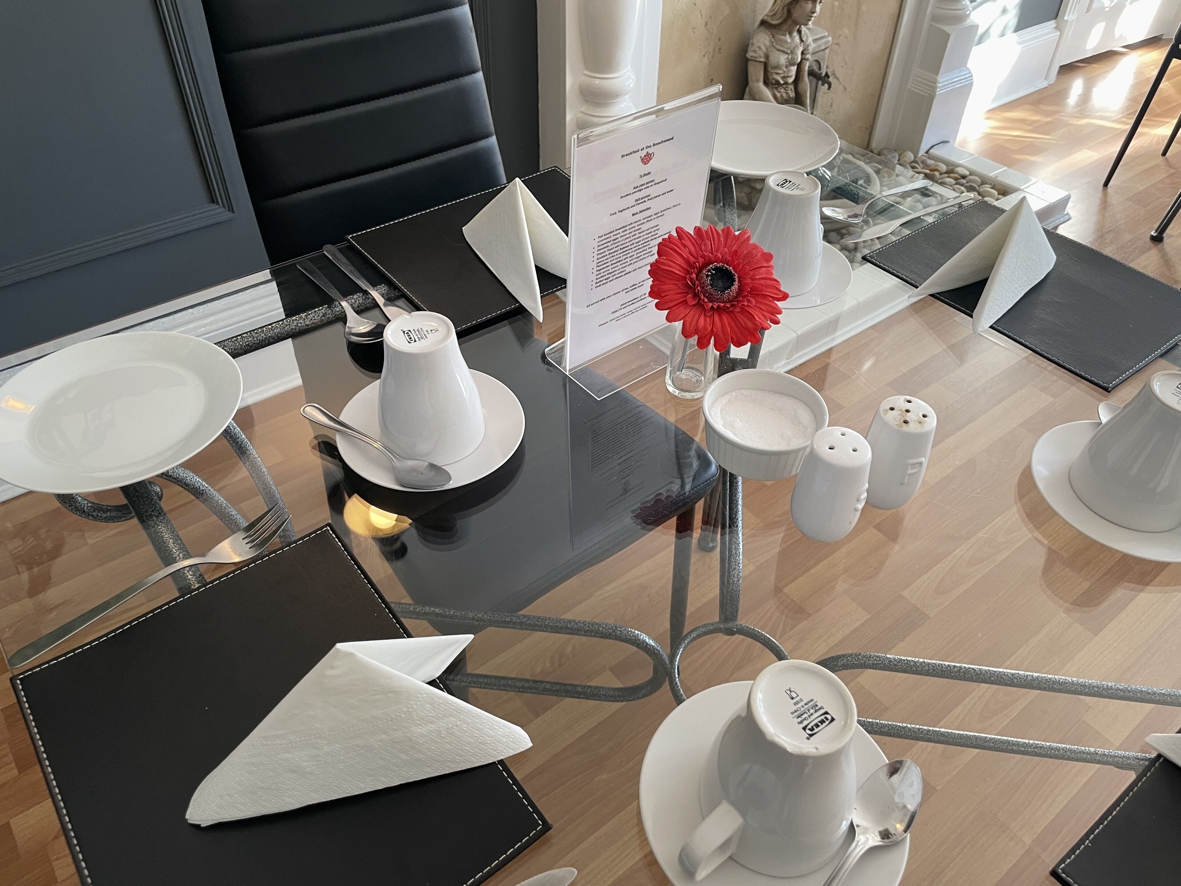 A dining area with a black table, white plates, and silverware. A menu sign is visible, and a red flower is placed on the table.