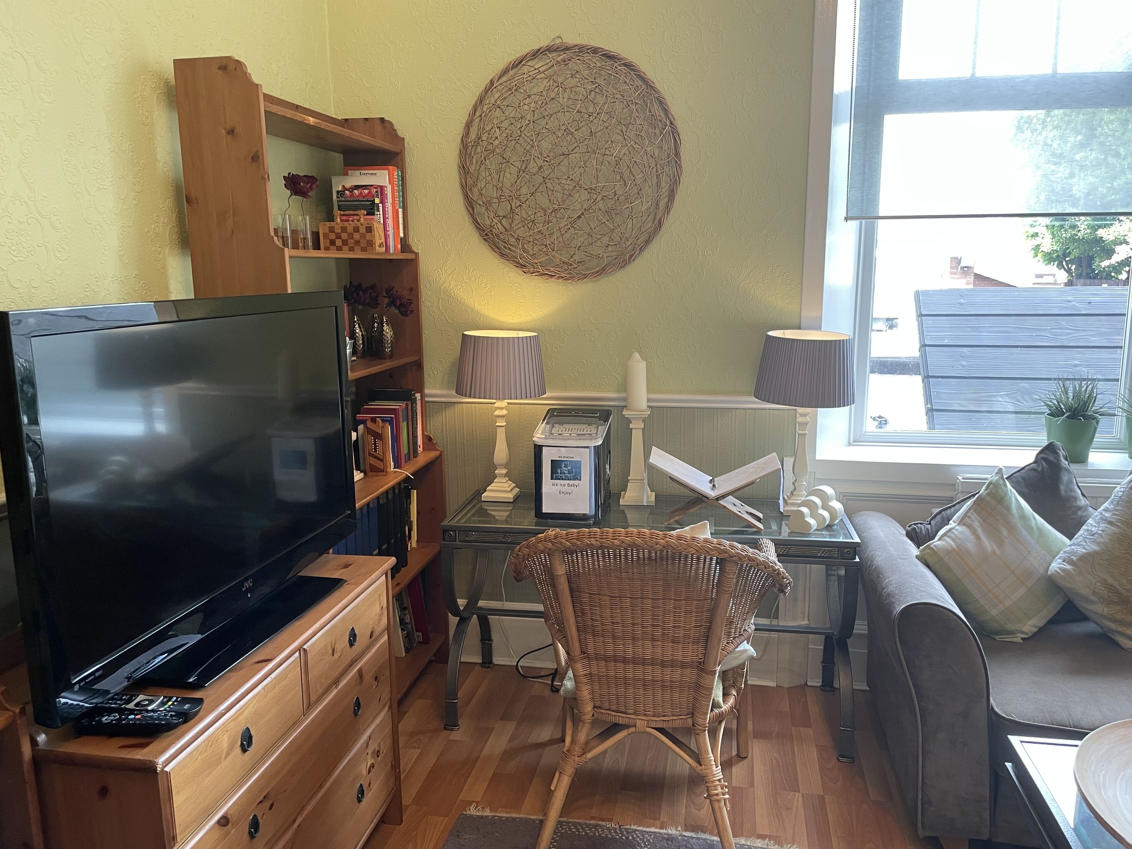 A cozy living room with a flat-screen TV, wooden furniture, a wicker chair, and a view of the outside through a window.
