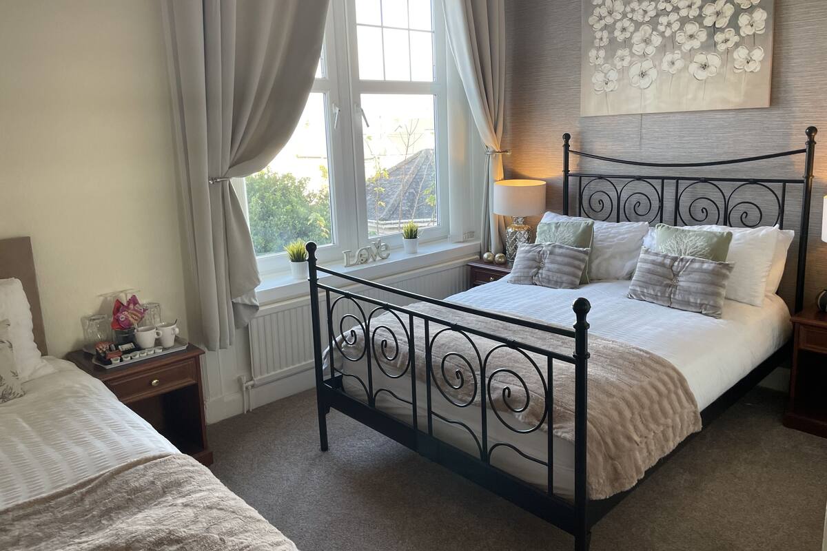 A bedroom with a bed, a window with curtains, a bedside table with a lamp, and a wall with a floral artwork.