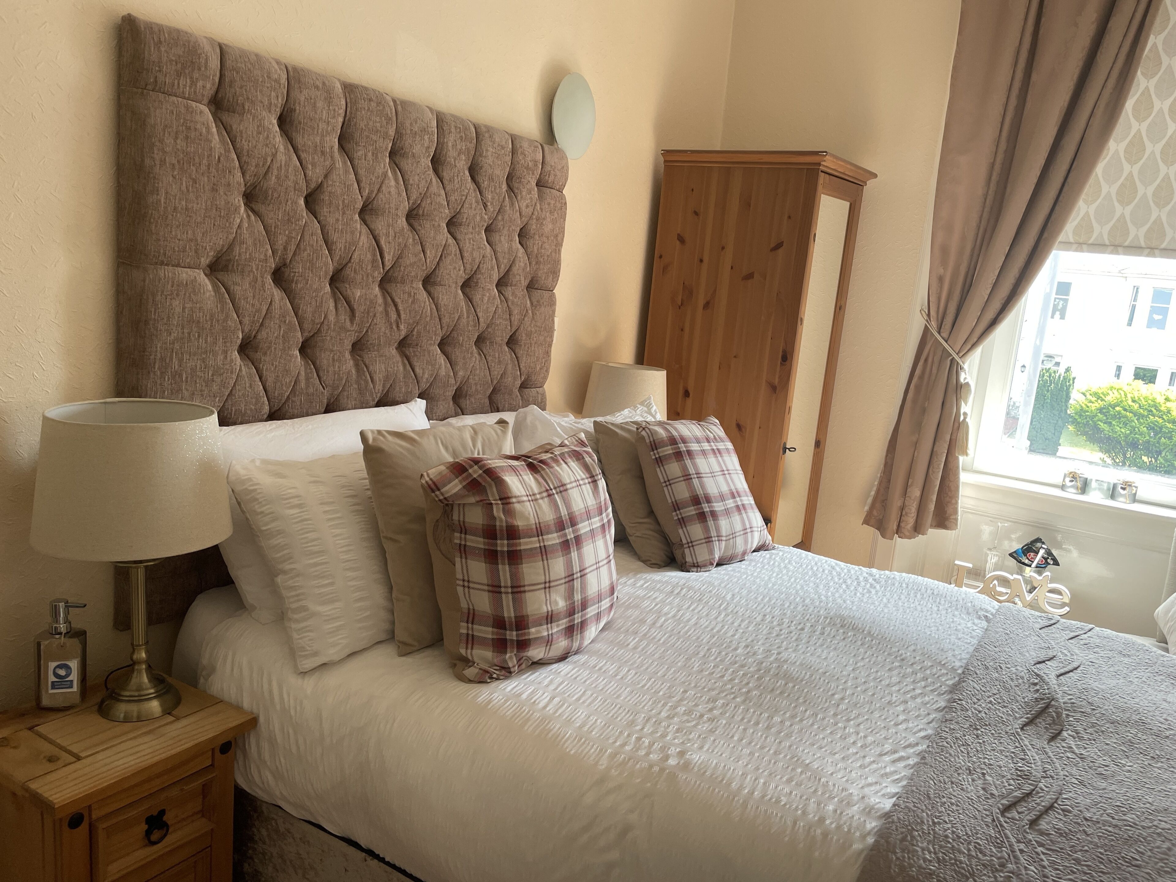 A bedroom with a tufted headboard, a wooden wardrobe, a bedside table with a lamp, and a window with curtains.